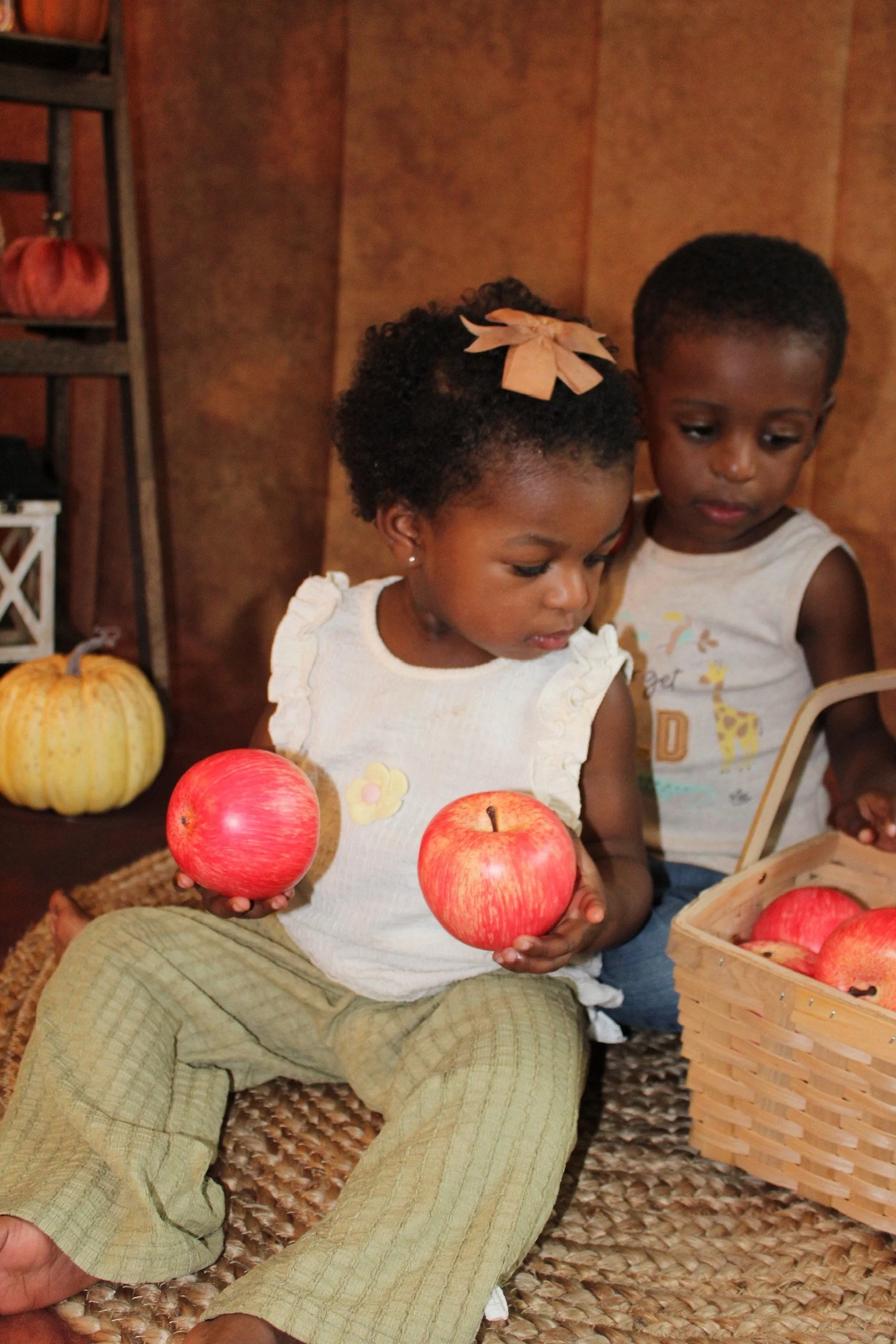 Two young children sitting on a woven mat holding red apples and a basket of apples, with pumpkins in the background.