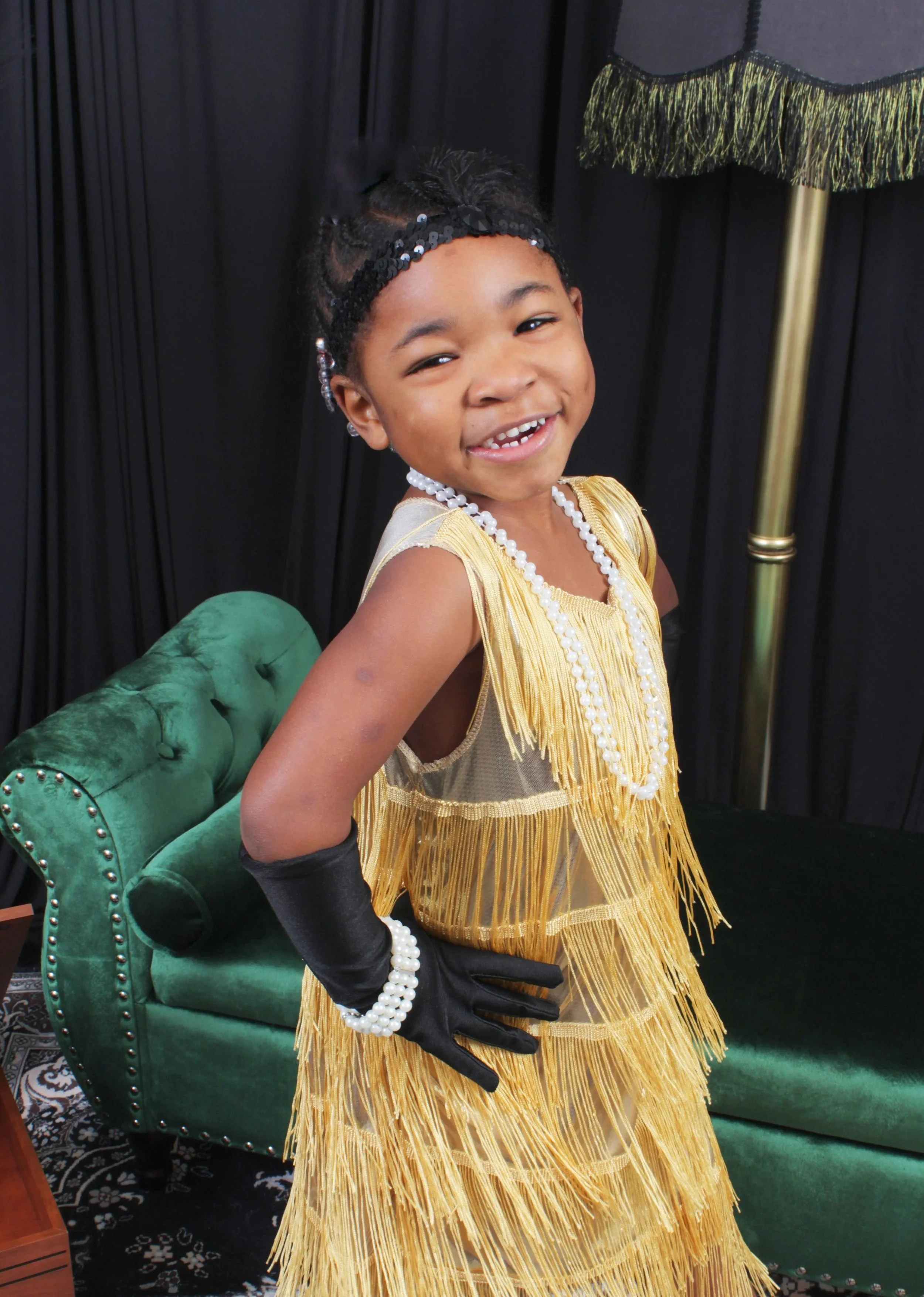 Young girl dressed in vintage gold flapper costume with fringe, pearl accessories, and black gloves, smiling and posing in front of a black curtain and green velvet armchair.