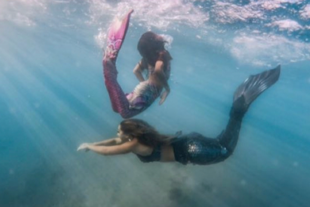 One child and a woman with mermaid tails swimming underwater, one with a pink tail and the other with a black tail.