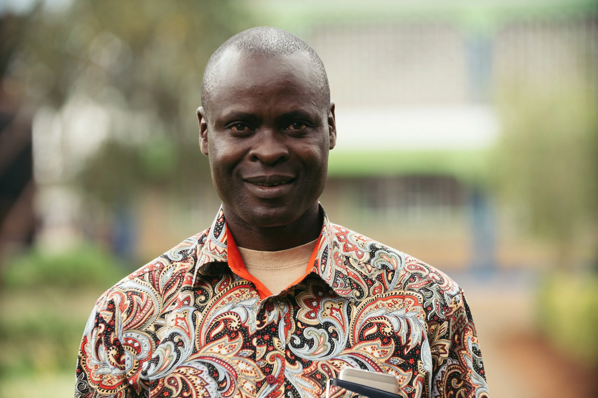 A portrait of a man with a serious expression, dark skin, and a shaved head, wearing a patterned shirt, set against a plain white background.