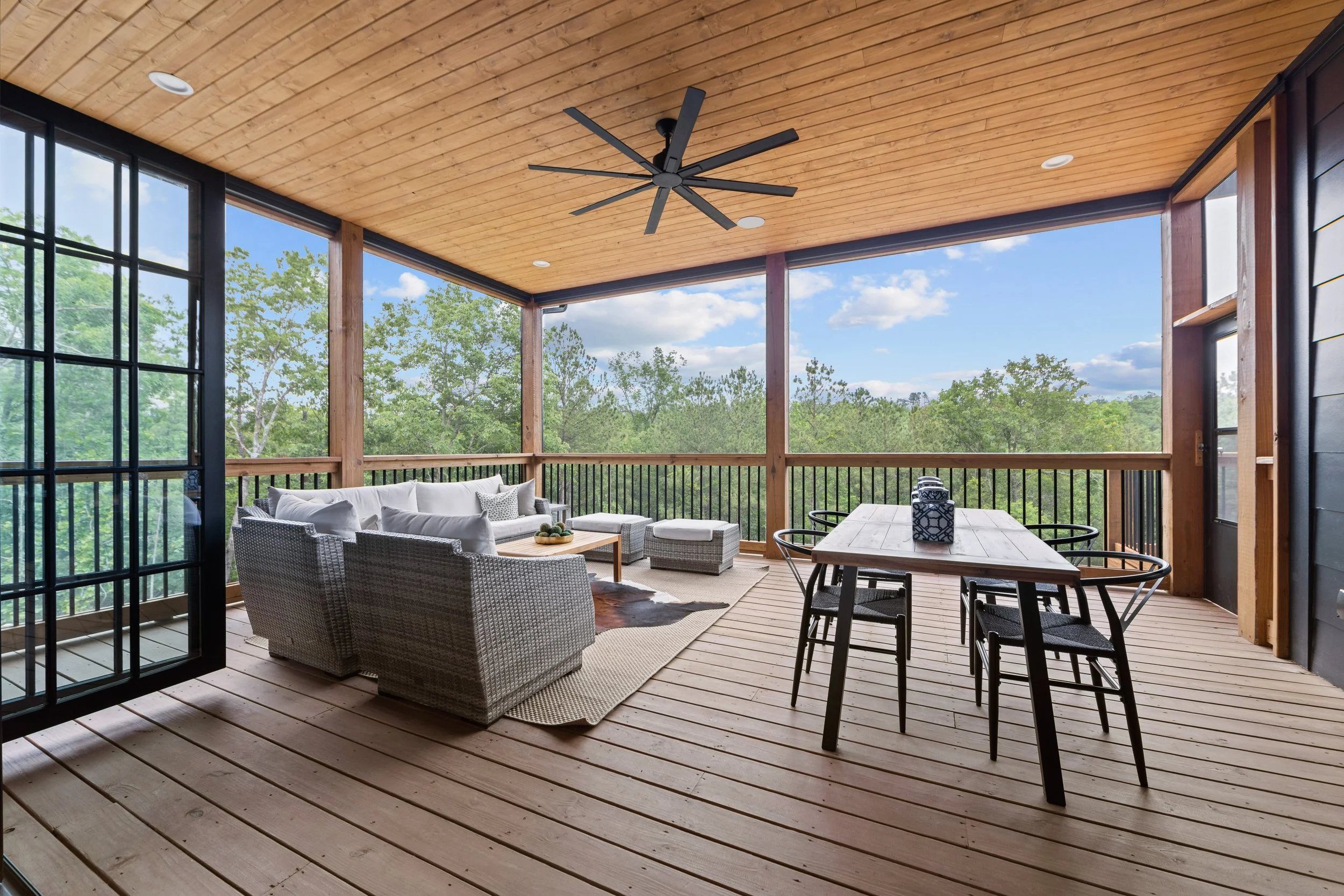Covered outdoor porch with wooden flooring and ceiling, black ceiling fan, wicker sofa with white cushions, wooden dining table with black chairs, surrounded by trees and blue sky.