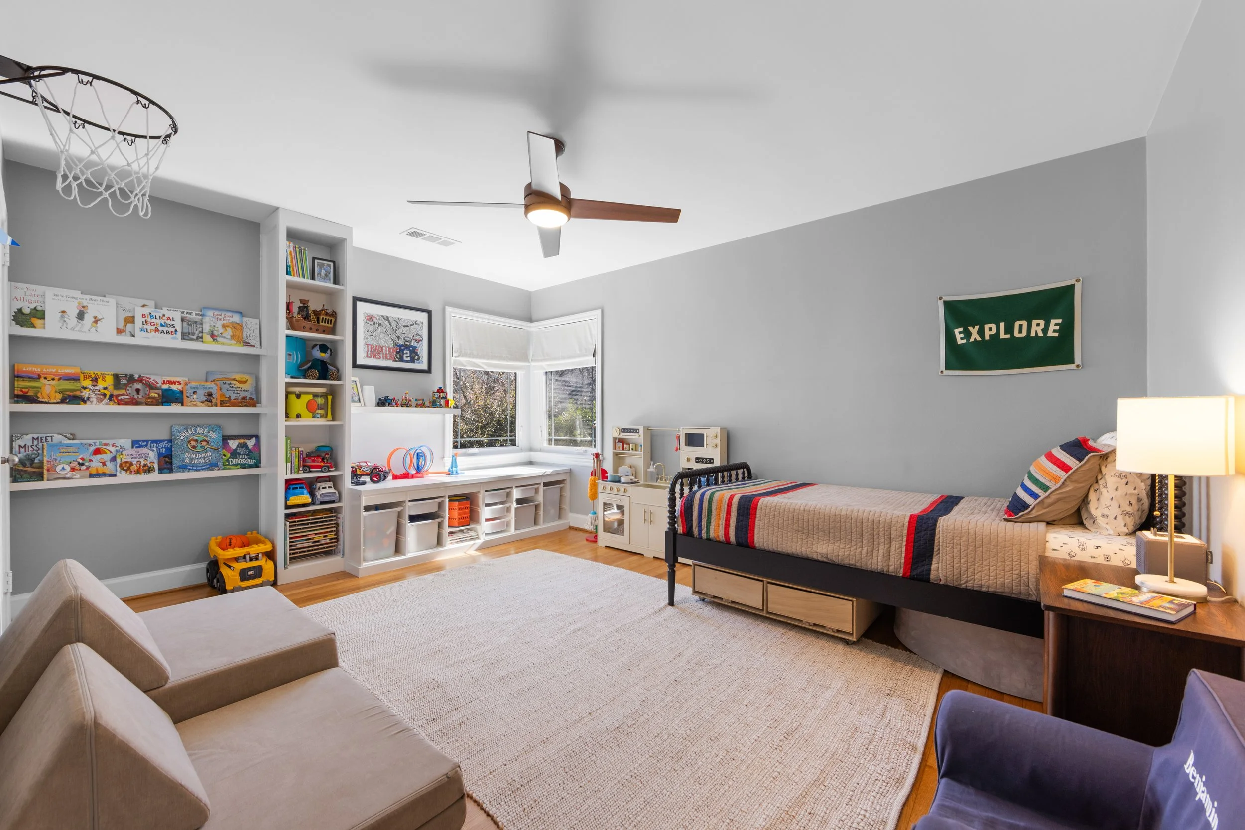 Child's bedroom with a bed, bookshelf filled with books and toys, beige armchair, and a rug. A window with blinds, a ceiling fan, and a wall sign reading 'EXPLORE' are visible.