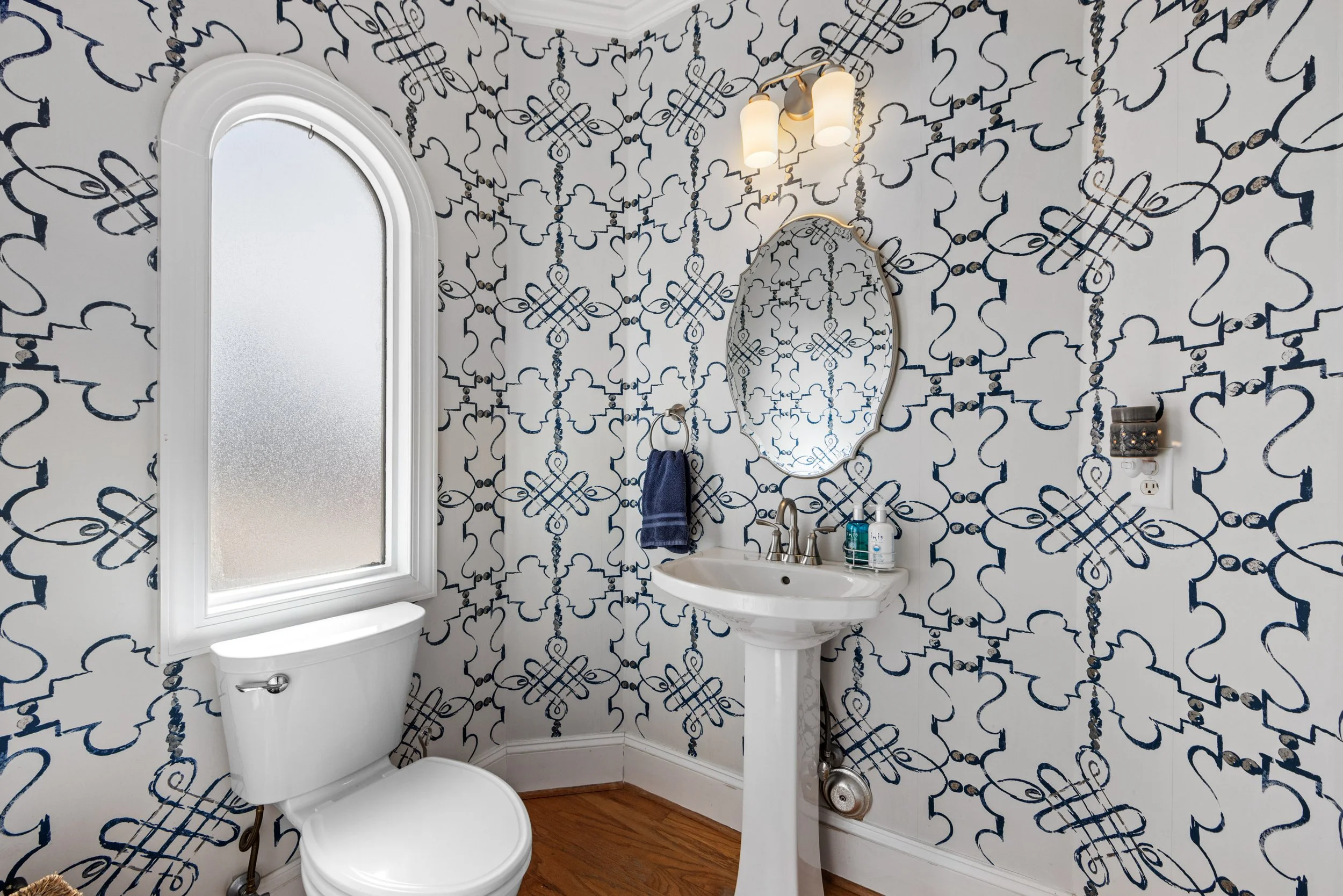 A small bathroom with patterned wallpaper featuring black designs on a white background, a white toilet, a white pedestal sink with a faucet, a mirror above the sink, a wall-mounted light fixture, a towel ring with a navy blue towel, and a frosted wi
