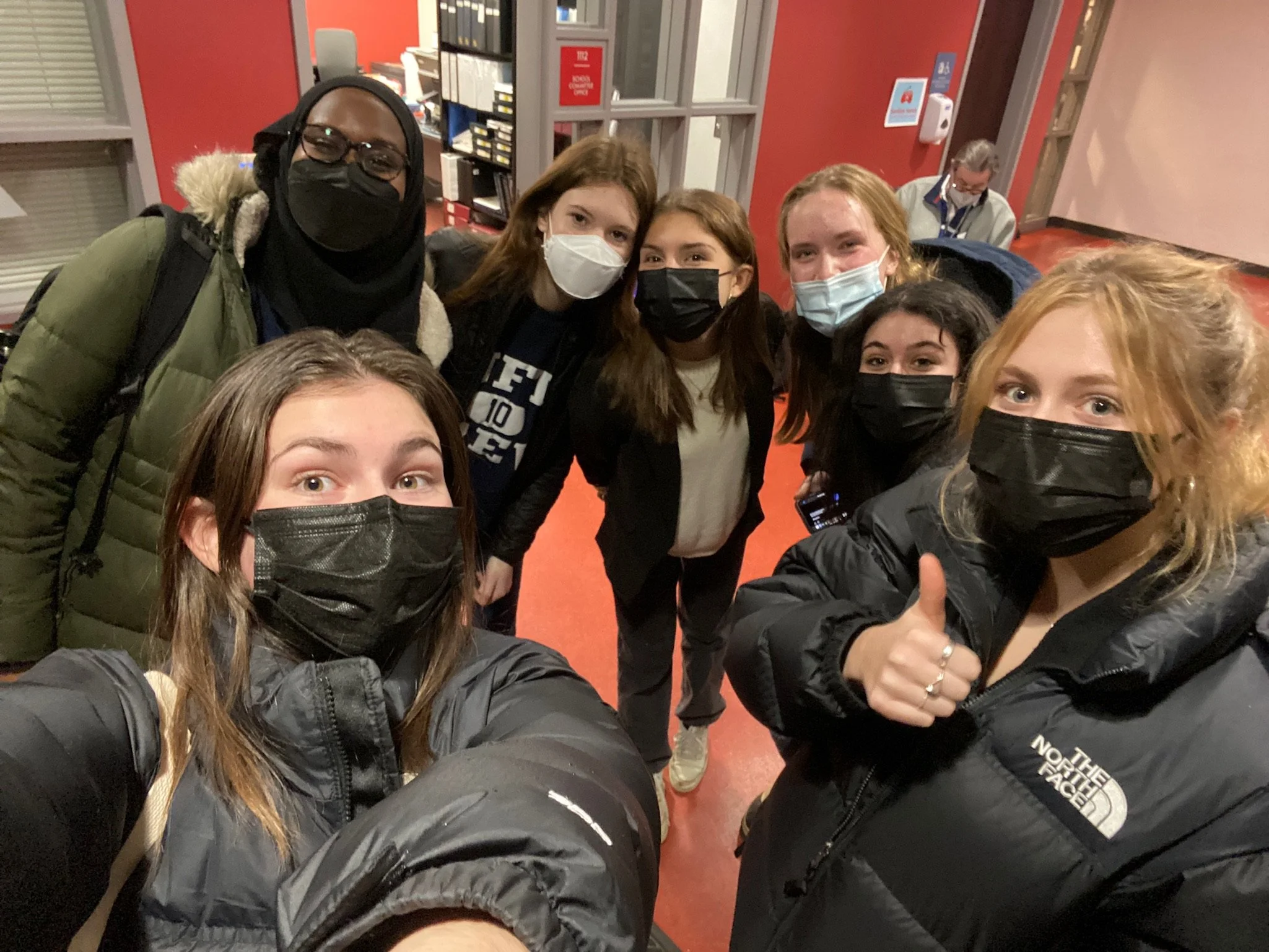 Group of people wearing masks indoors, dressed in winter clothing, posing for a selfie.