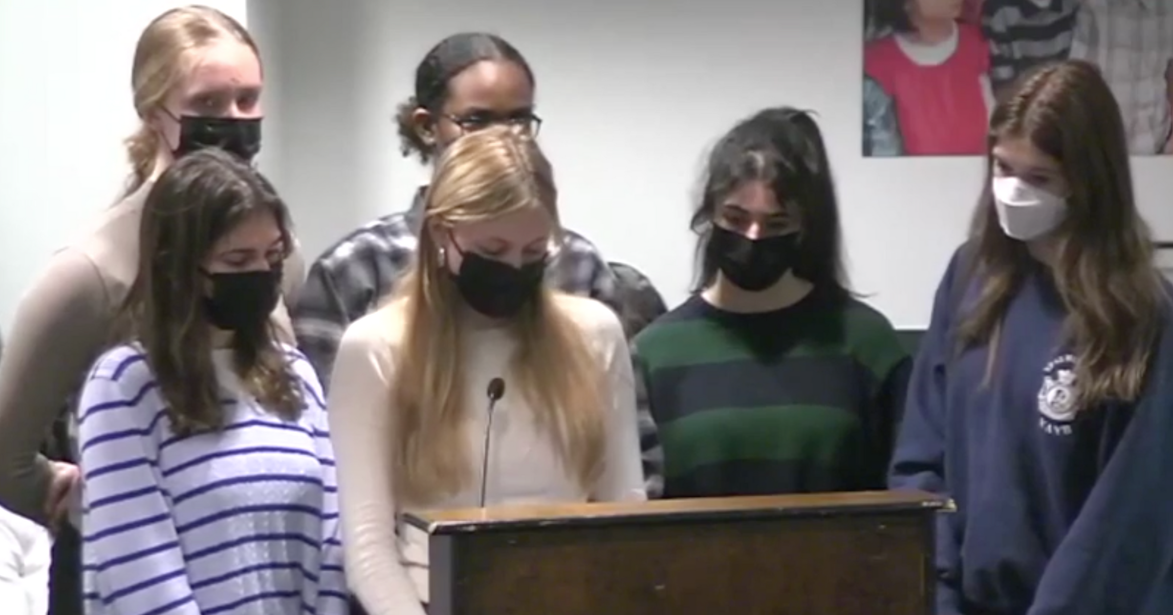 A group of six people standing together, all wearing face masks, in front of a podium indoors.