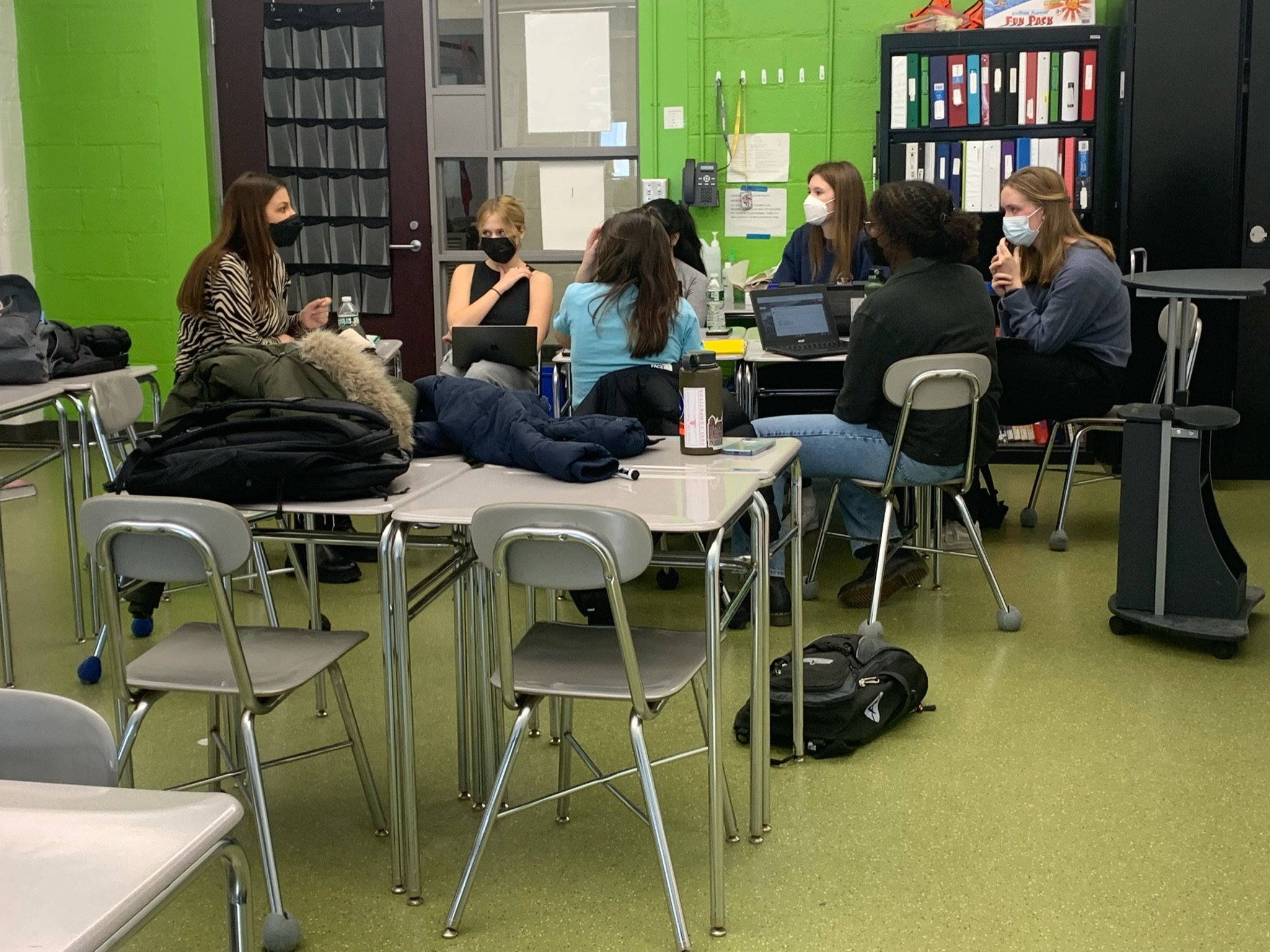 A group of people wearing masks seated around a table in a classroom, engaged in discussion, with laptops and notebooks visible.