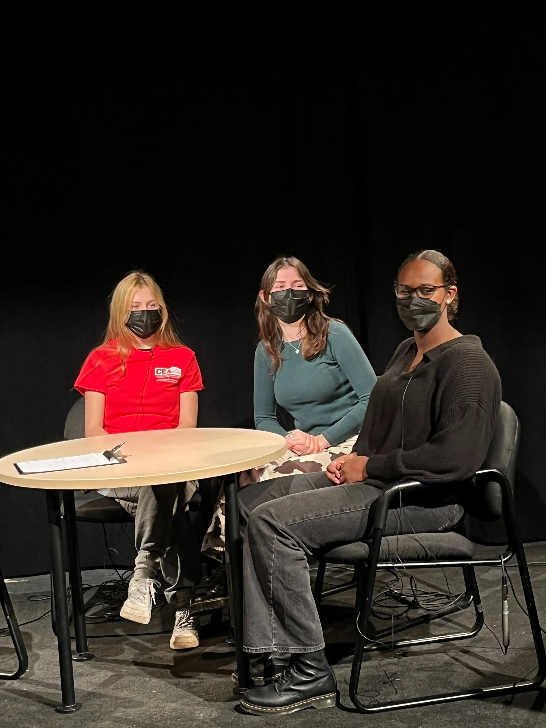 Three individuals wearing masks sitting at a round table on stage, with black background and visible wires.