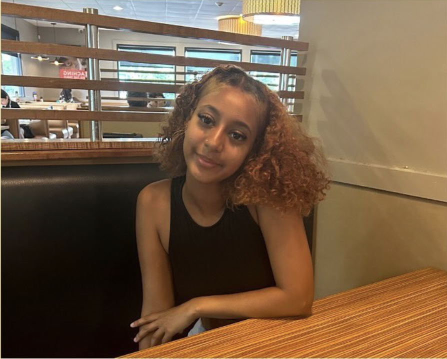 Person with curly hair sitting in a restaurant booth, wearing a black sleeveless top.
