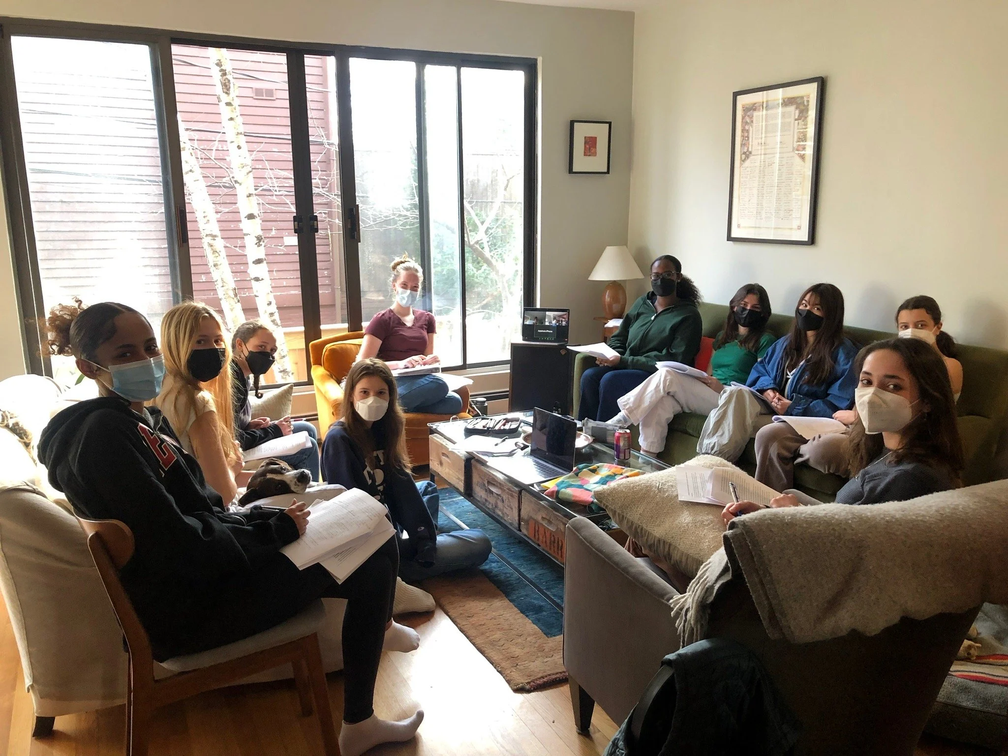 Group of people wearing masks sitting in a living room, engaged in a meeting or study session, with papers and a laptop on the table.