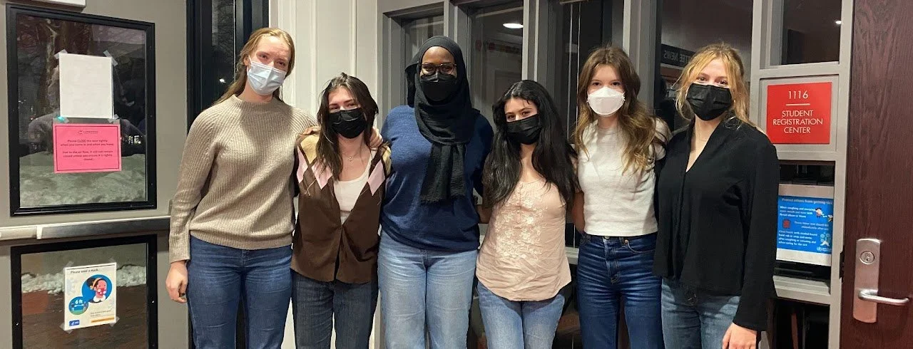 Group of six women wearing masks standing indoors in front of a window.