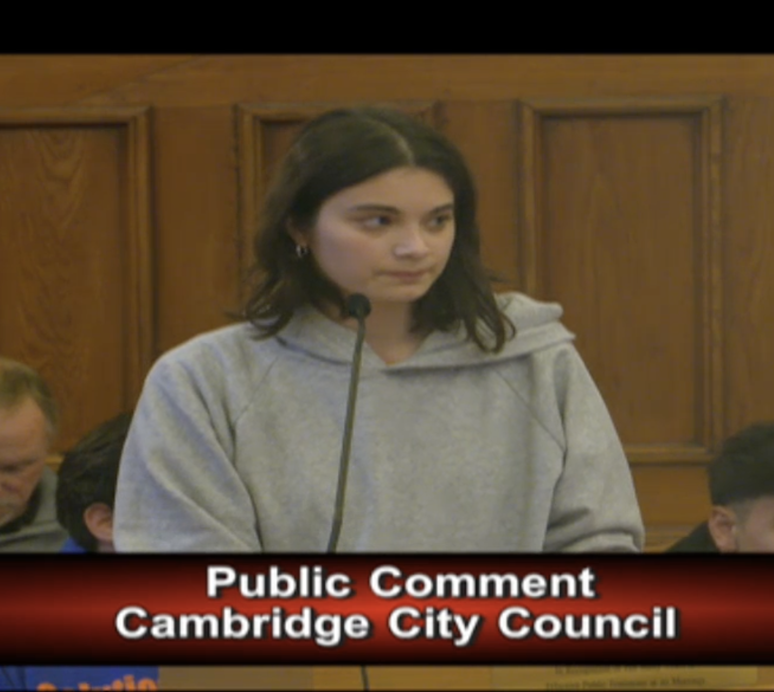 A person in a gray hoodie speaking at a microphone during a Cambridge City Council public comment session.