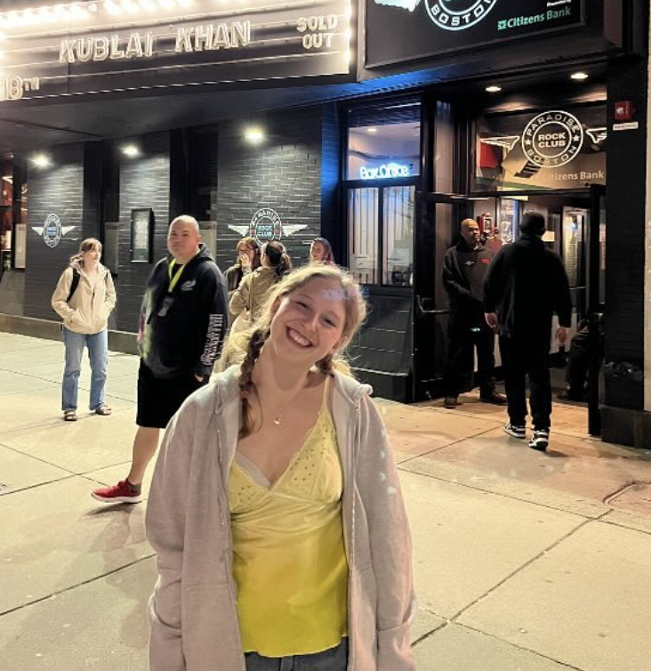 A woman smiling outside the Paradise Rock Club in Boston, with a sign showing 'Kublai Khan' sold out. People are lined up near the entrance, and the club's neon sign is visible.