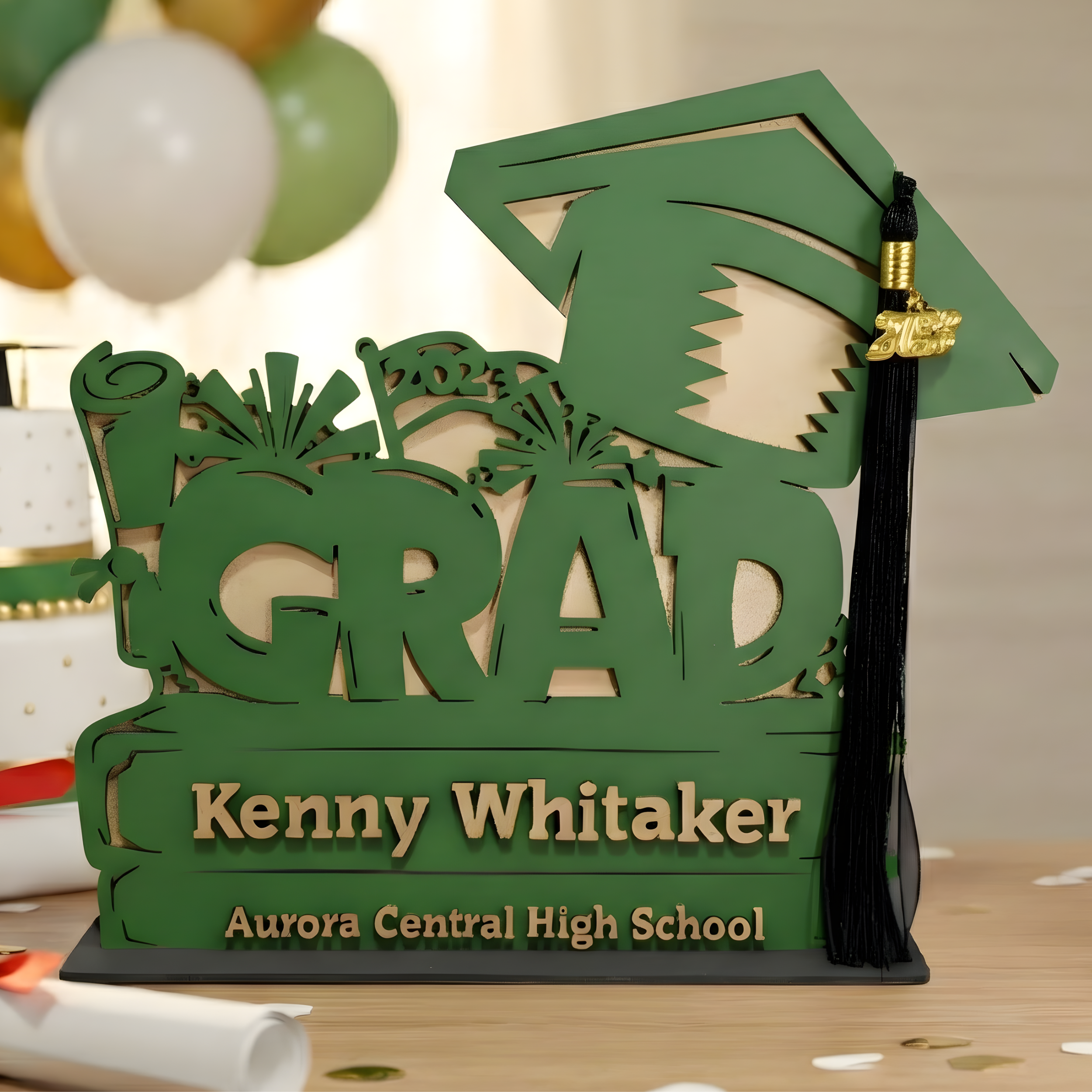 Personalized green "GRAD" sign with name "Kenny Whitaker" holding a black and gold graduation tassel.
