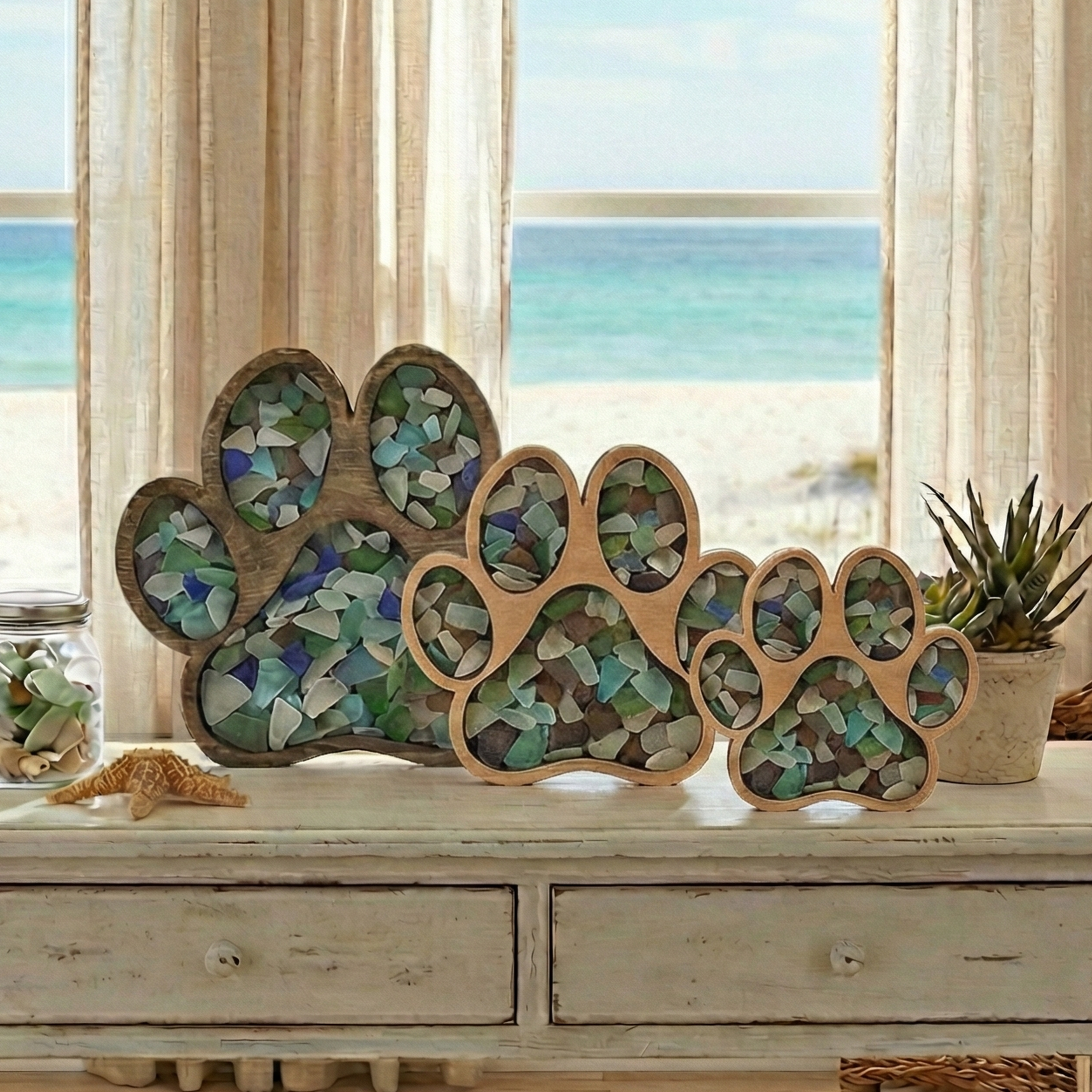 Three tiered wooden paw-print displays in various sizes, each filled with a collection of colorful sea glass pieces, arranged on a white console table against a bright beach house background.