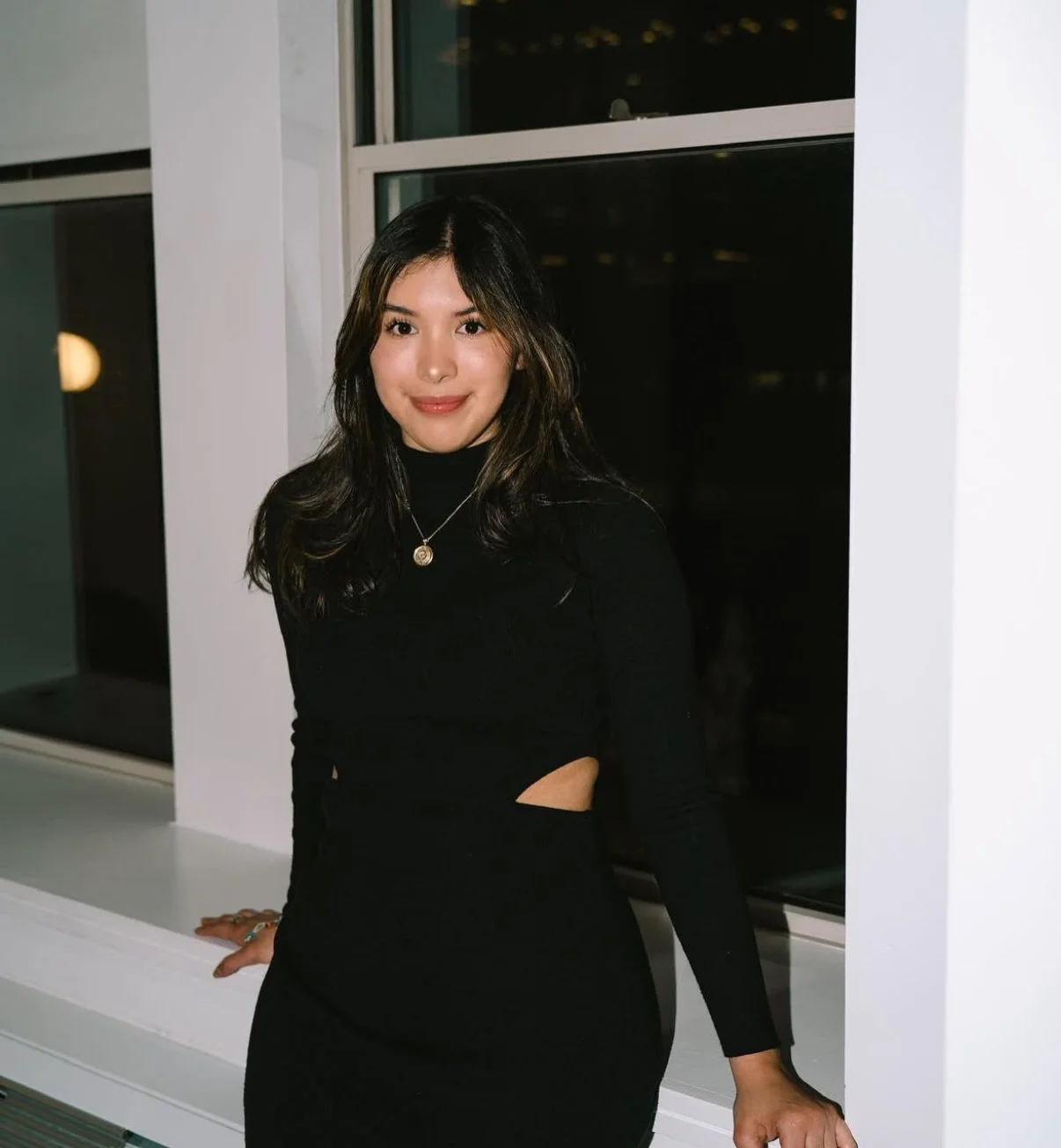 Woman in black dress standing by a window at night.