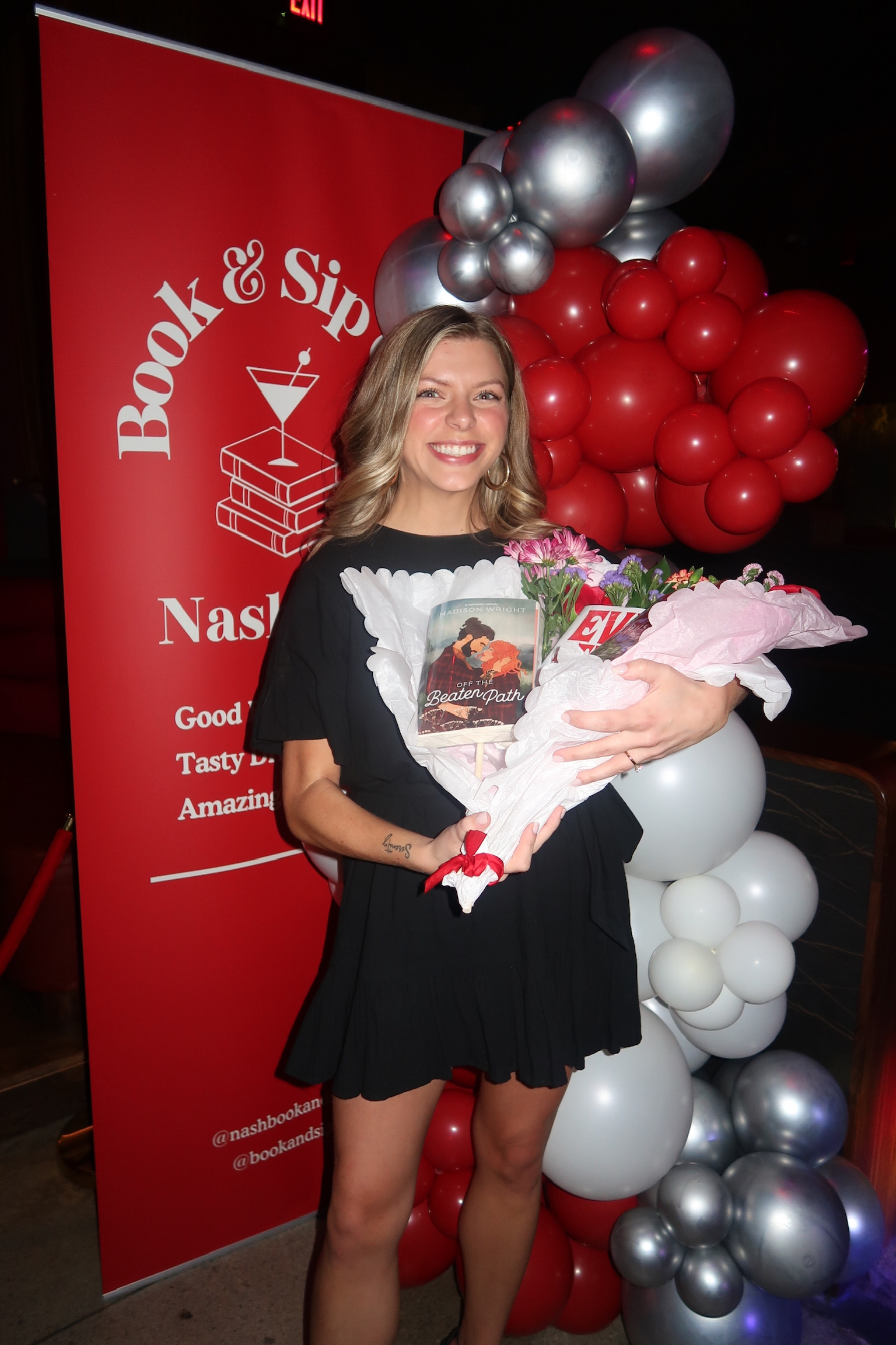A woman in a white dress standing next to a red sign for "Book & Sip Club" in Nashville. The sign mentions "Good Books, Tasty Drinks, Amazing Company."
