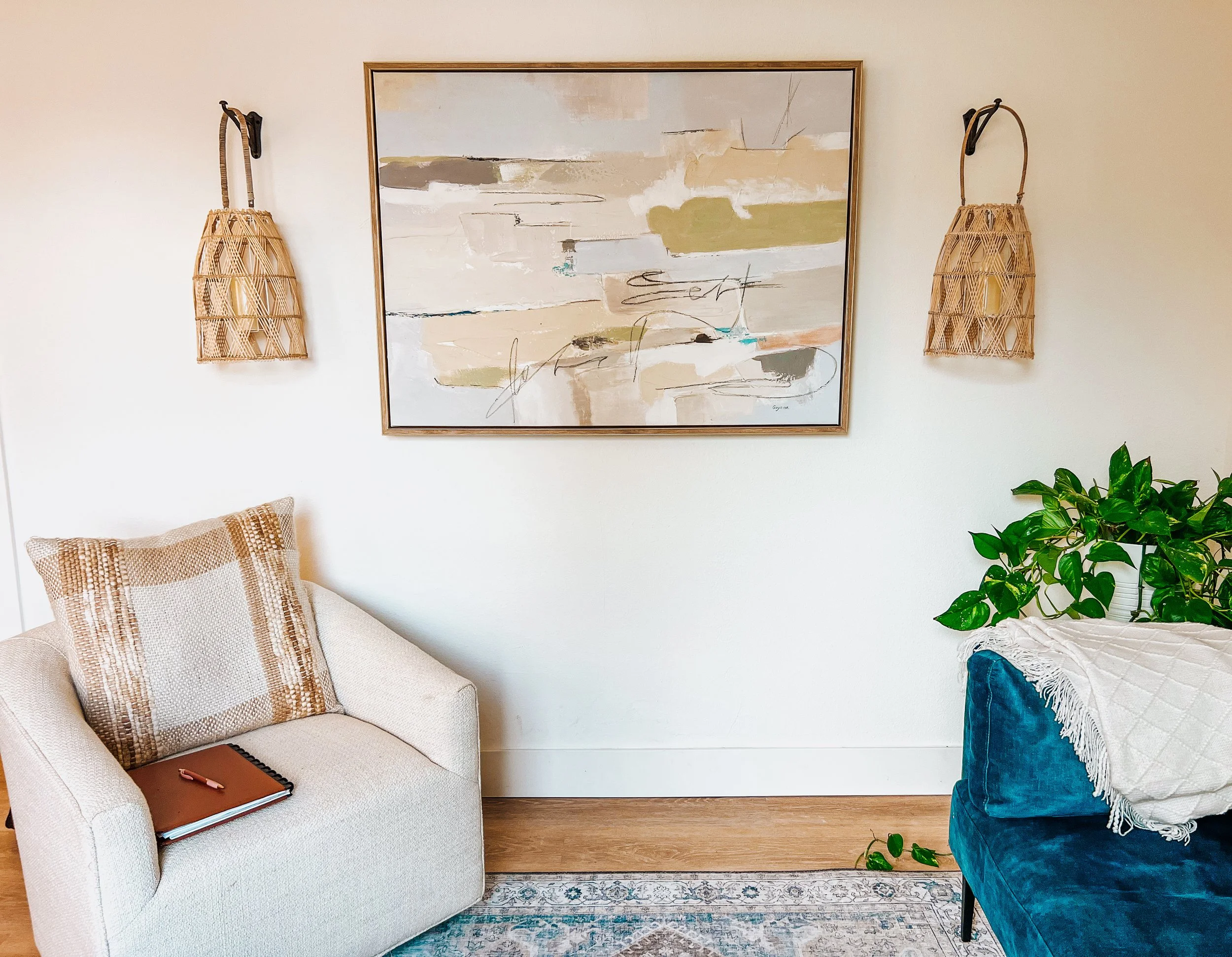 Living room with abstract art, beige armchair, blue sofa, hanging rattan lights, and a potted plant.