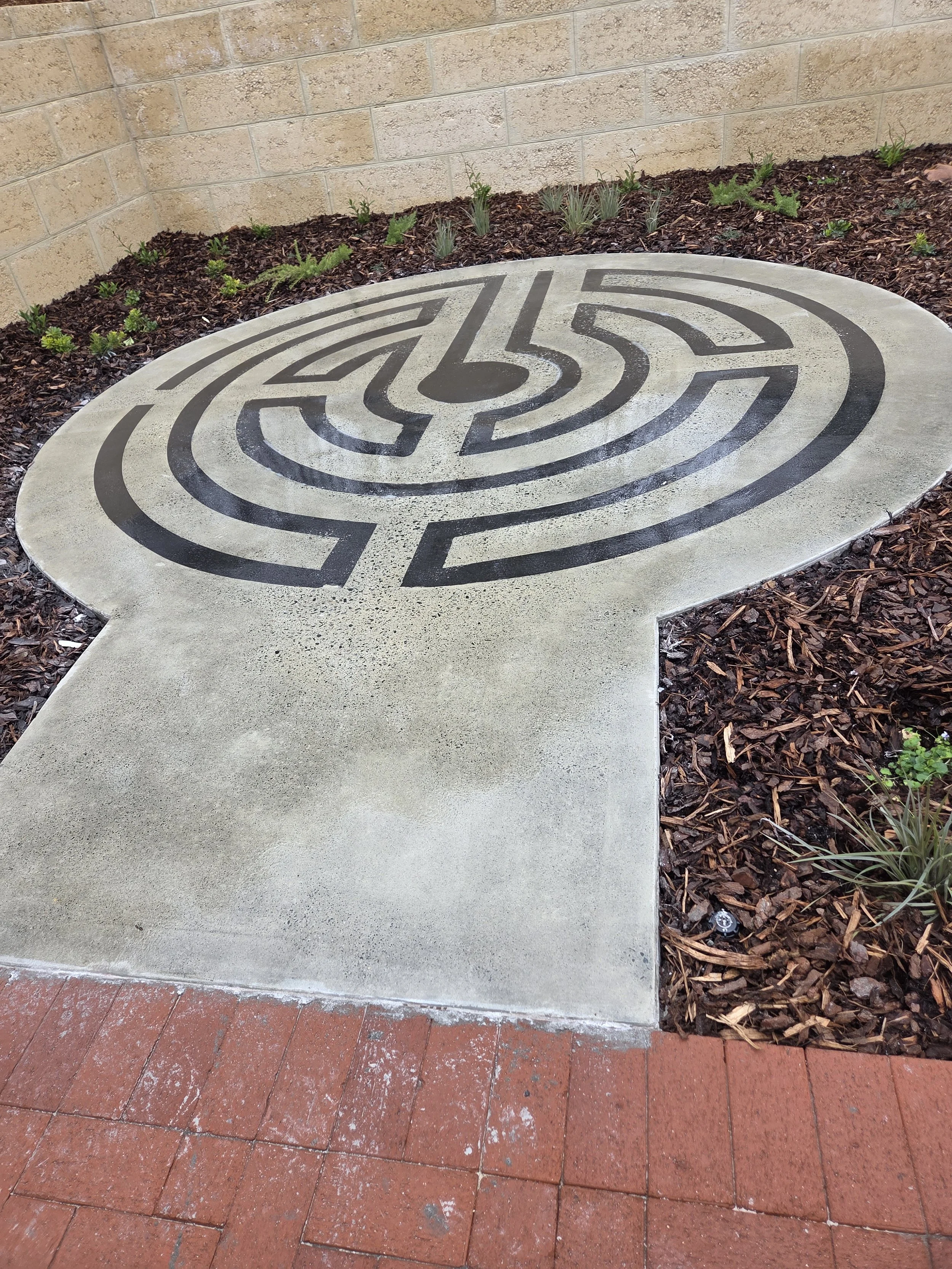 Concrete pathway with a maze design embedded in it, surrounded by mulch and small plants, with a brick sidewalk in the foreground and a brick wall in the background.