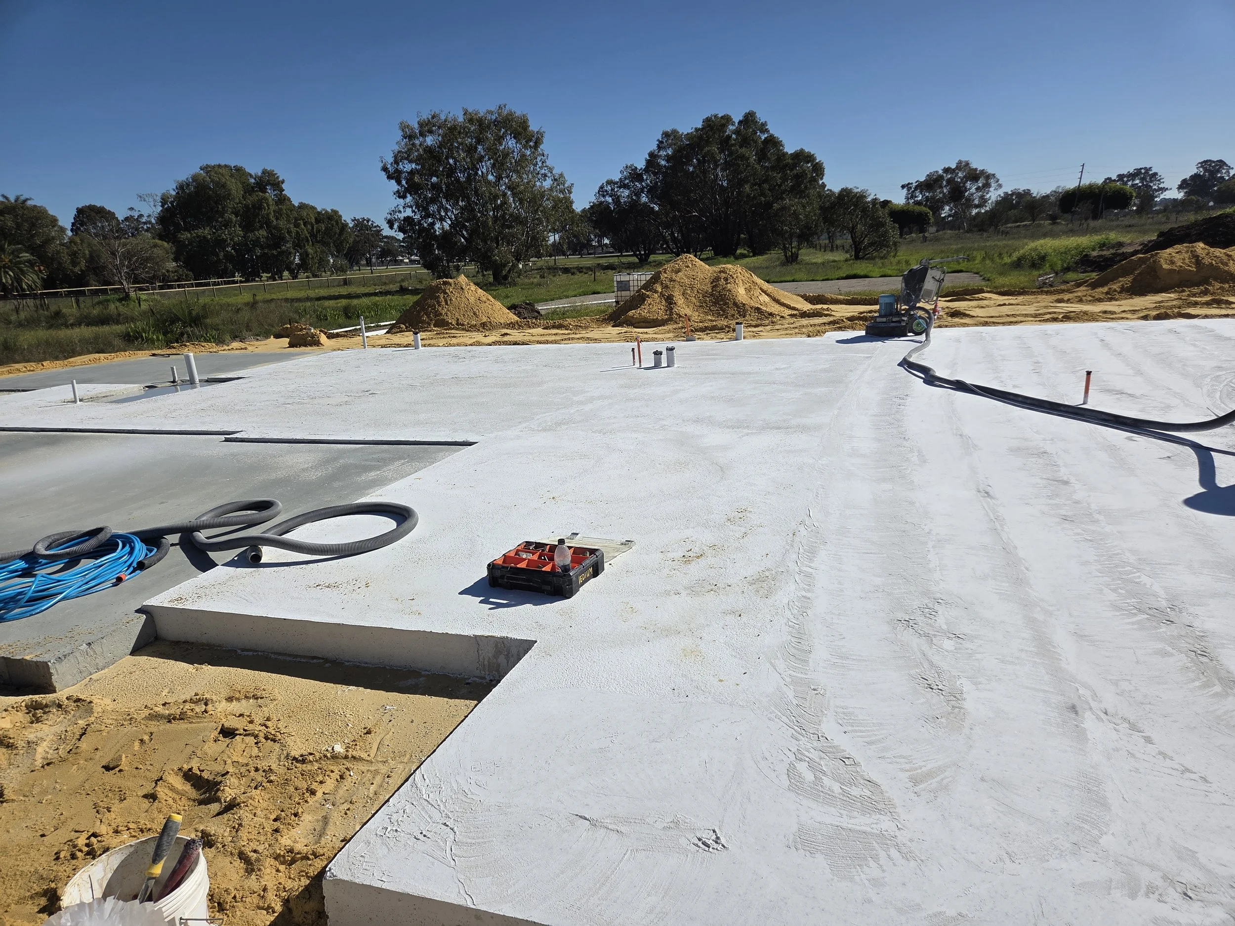 Construction site with a freshly poured concrete slab, equipment, and surrounding dirt mounds, with trees and blue sky in the background.