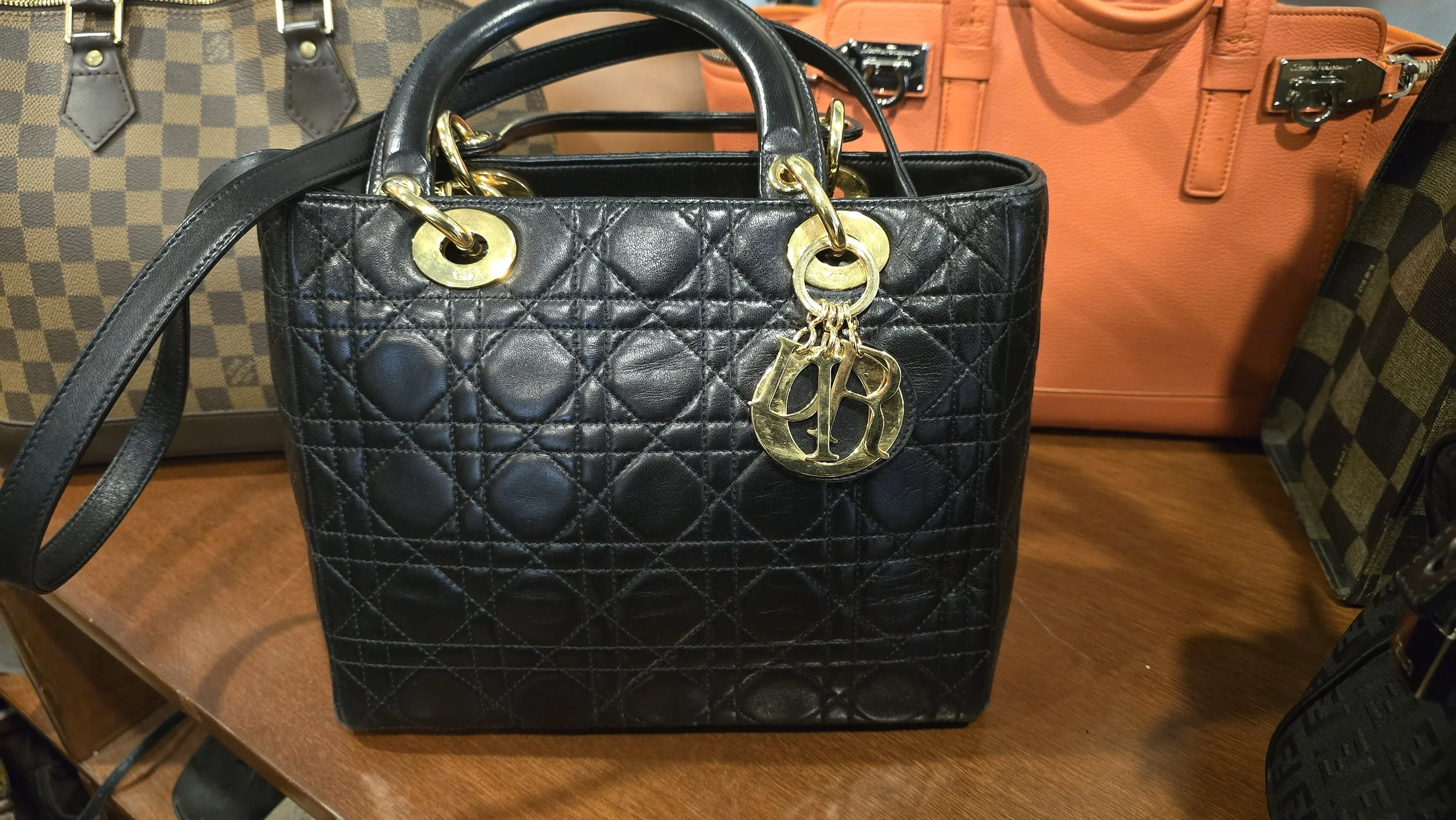 Black quilted leather handbag with gold hardware and attached gold letter charms on display with bouclé and orange designer handbags in the background.