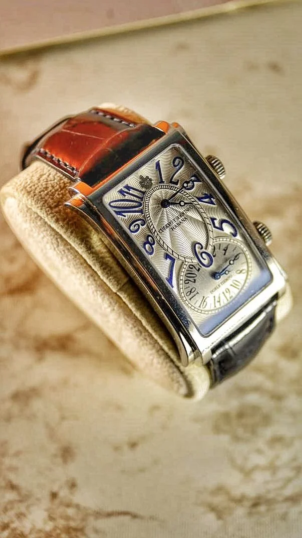 A luxury wristwatch with a rectangular face, silver dial, blue numerals, and leather strap, resting on a beige cushion.
