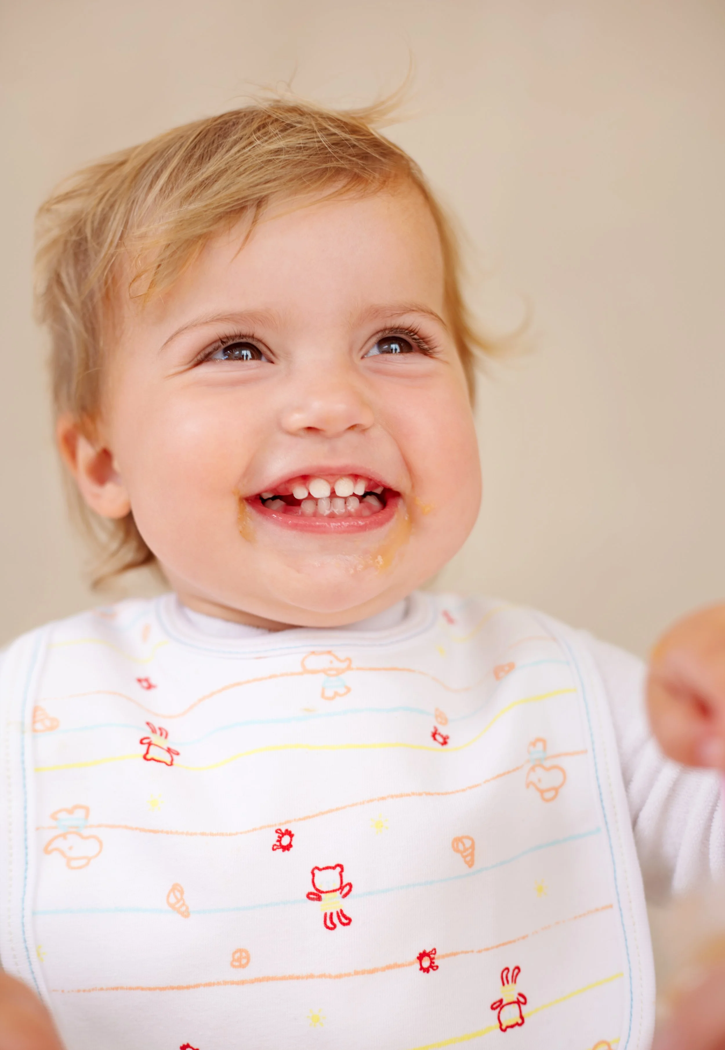 Close-up of a smiling toddler with messy blonde hair, wearing a white shirt with colorful animal and flower prints.