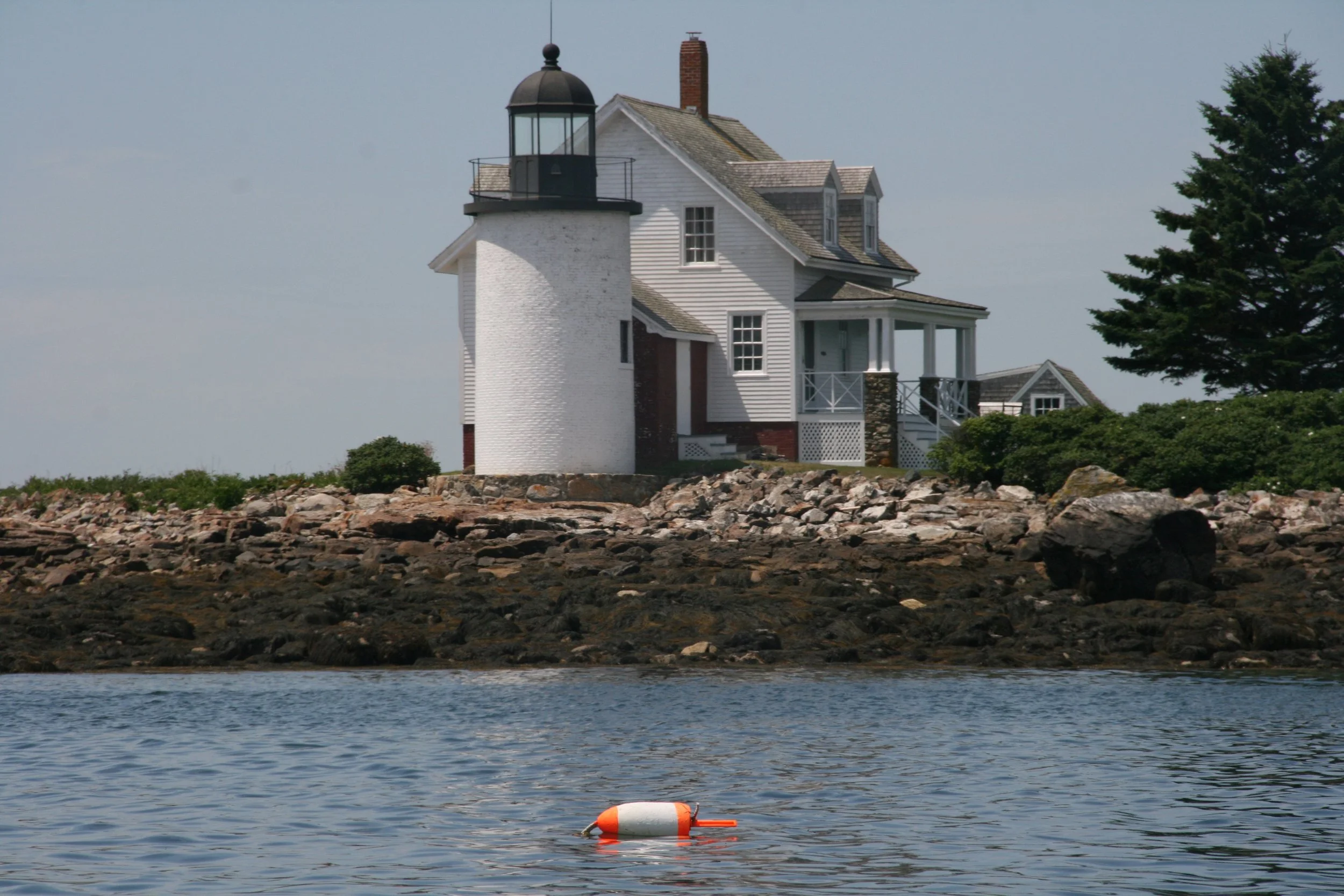 Lighthouse by a rocky shore with a white building and a buoy floating on the water in the foreground, under a clear sky.