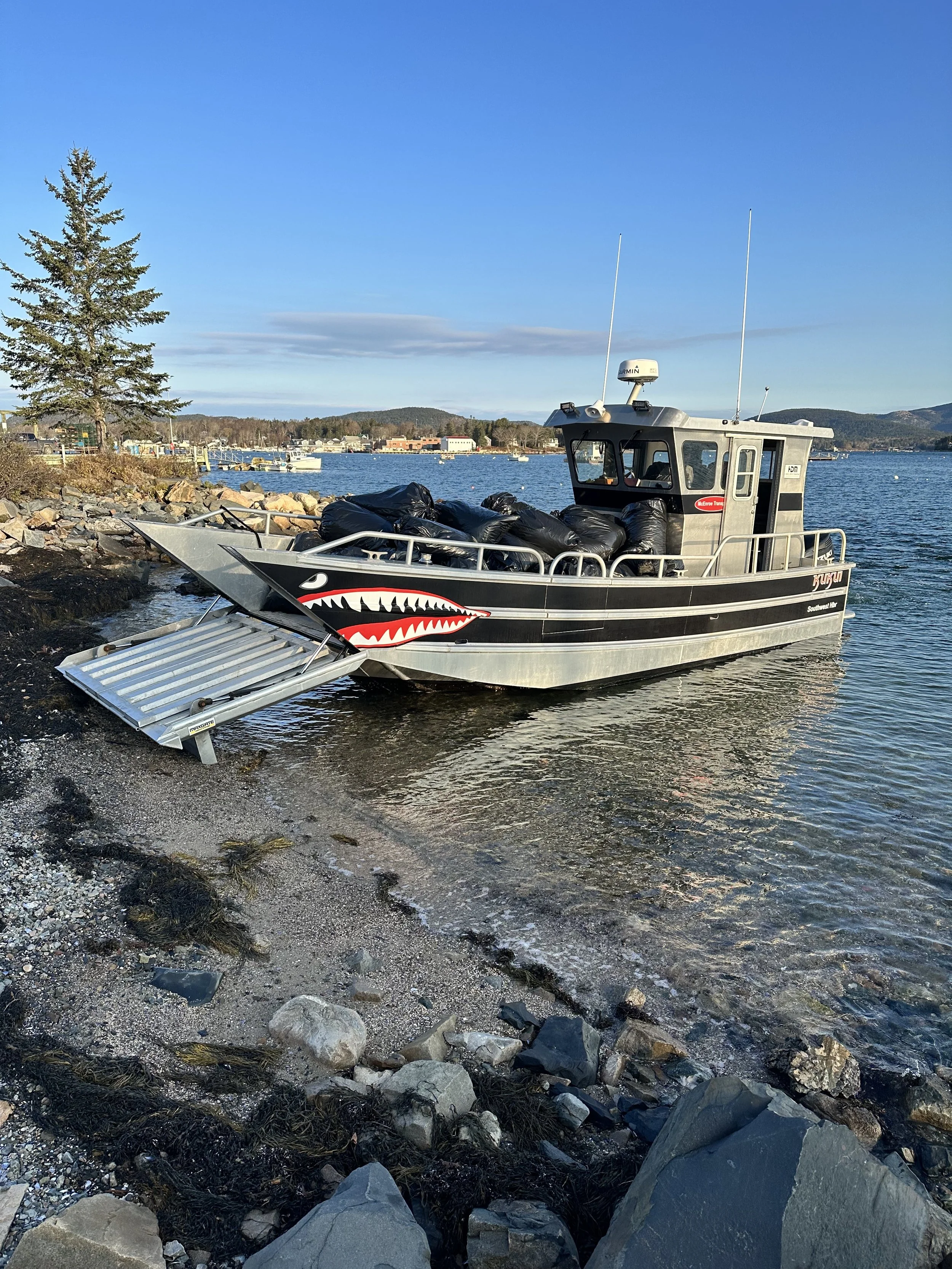 A boat with a painted shark face docked on a rocky shoreline, with a ramp lowered onto the sand. Several black bags are on the boat, and there's a background view of calm water, distant hills, and a clear blue sky.