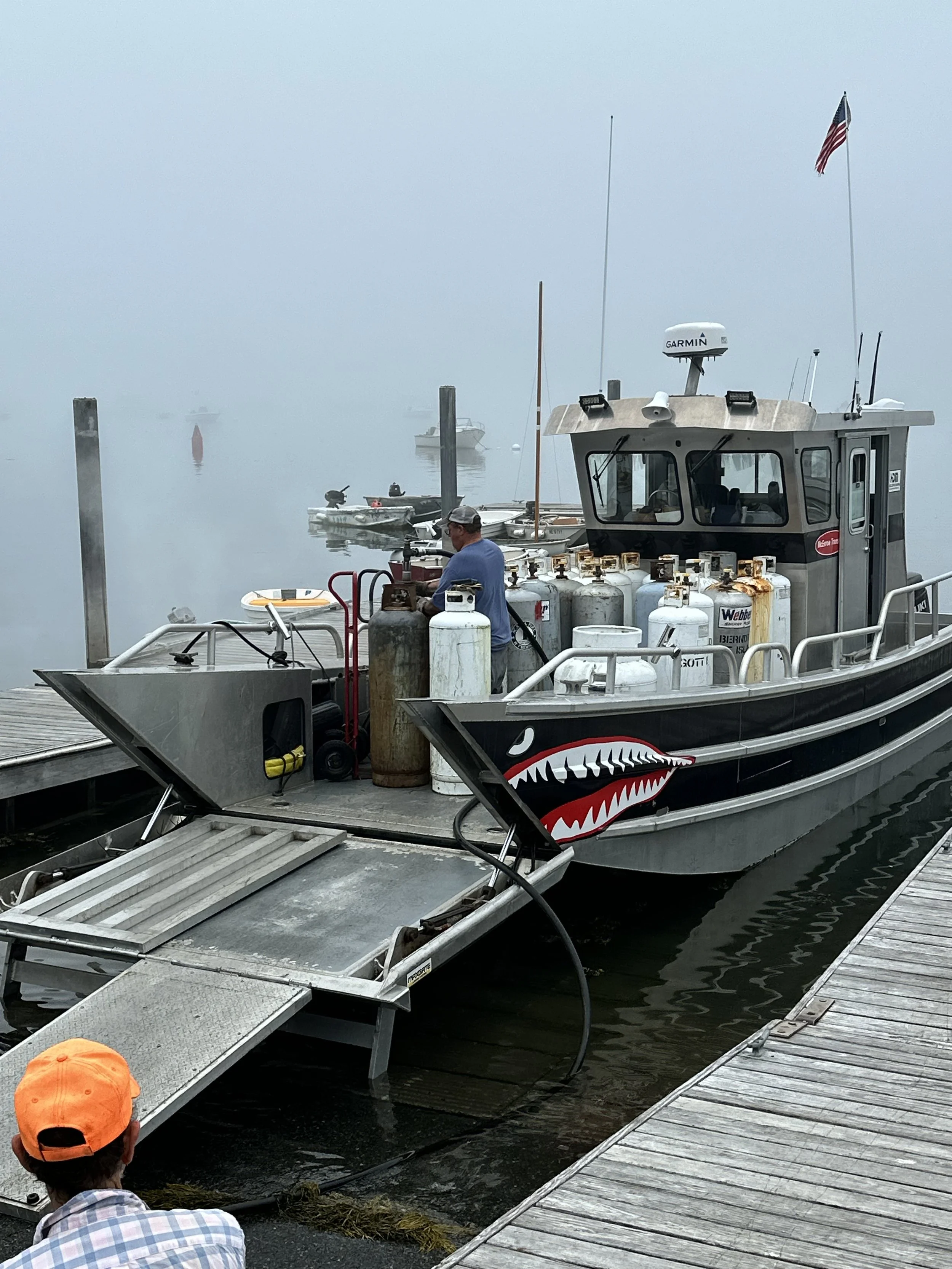 A metal boat docked at a pier, featuring a shark mouth design on its side. Several propane tanks are on board. A person in an orange cap is seated on the dock in the foreground, and American flag is on the boat. The background is foggy with a few boats visible in the mist.