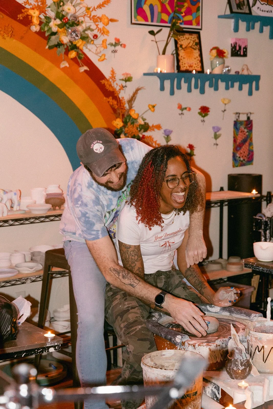 A couple sitting together at a pottery wheel making pottery and laughing