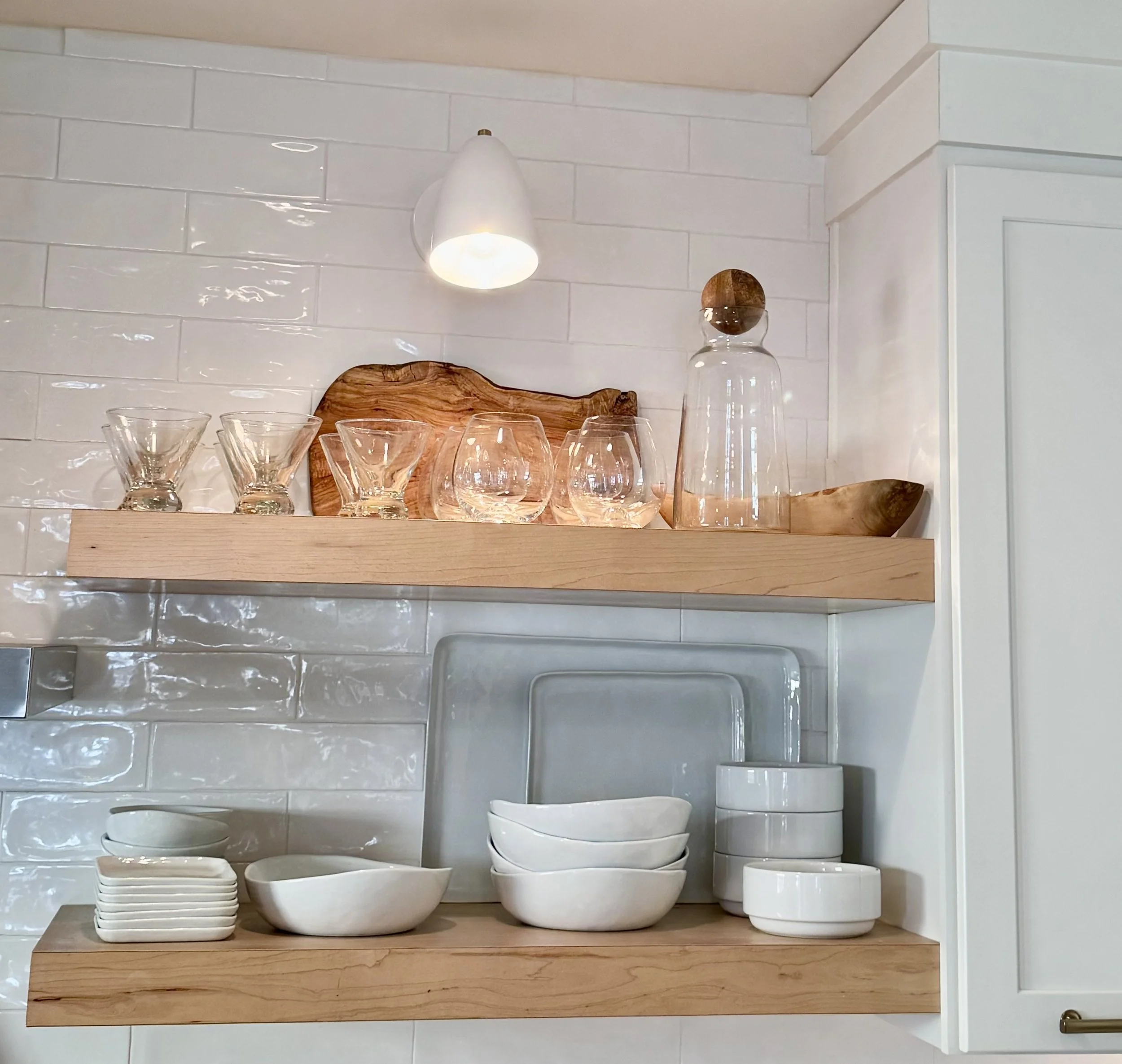 Open kitchen shelves in Kansas City with glassware, wooden cutting board, and ceramic bowls and dishes arranged on two wooden shelves against a white tiled wall.
