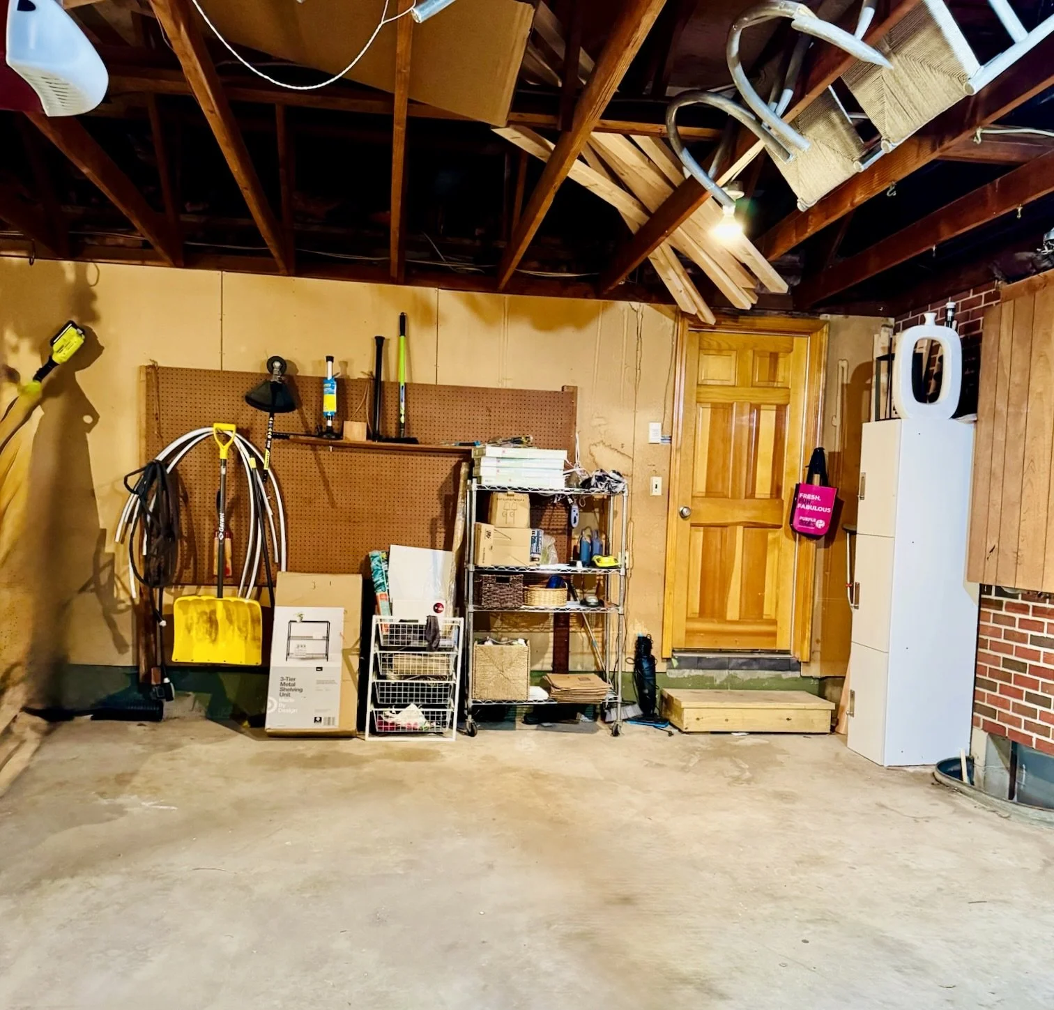 Garage interior with hanging gardening tools, wooden shelves filled with various items, and a wooden door. The garage floor is concrete, and the ceiling has exposed wooden beams and insulation.