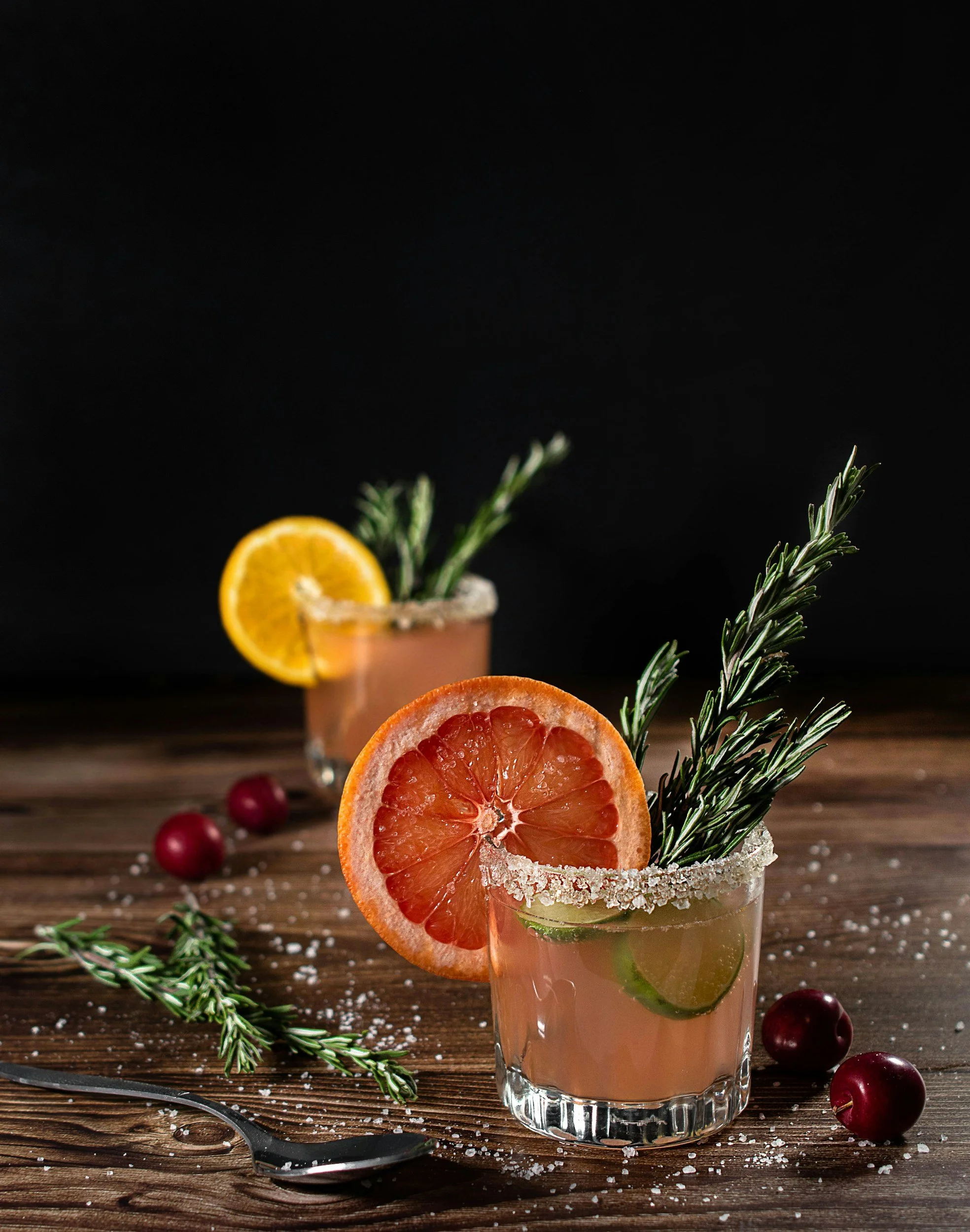 Two glasses of pink cocktail garnished with orange, lemon, lime slices, and rosemary sprigs on a wooden table, with cherries, salt, and a spoon nearby, against a dark background.