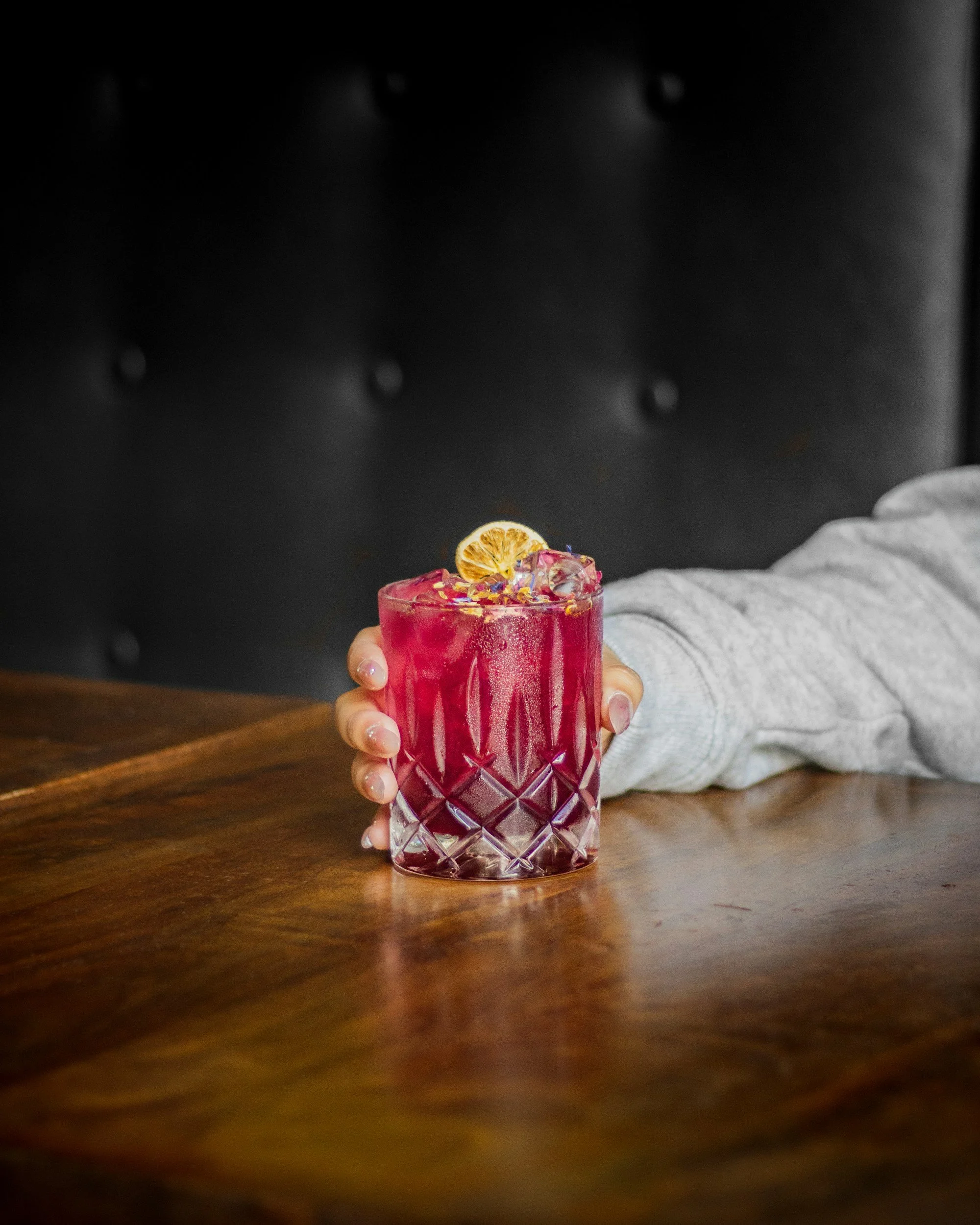 A person holding a pink cocktail garnished with a dried citrus slice on a wooden table.