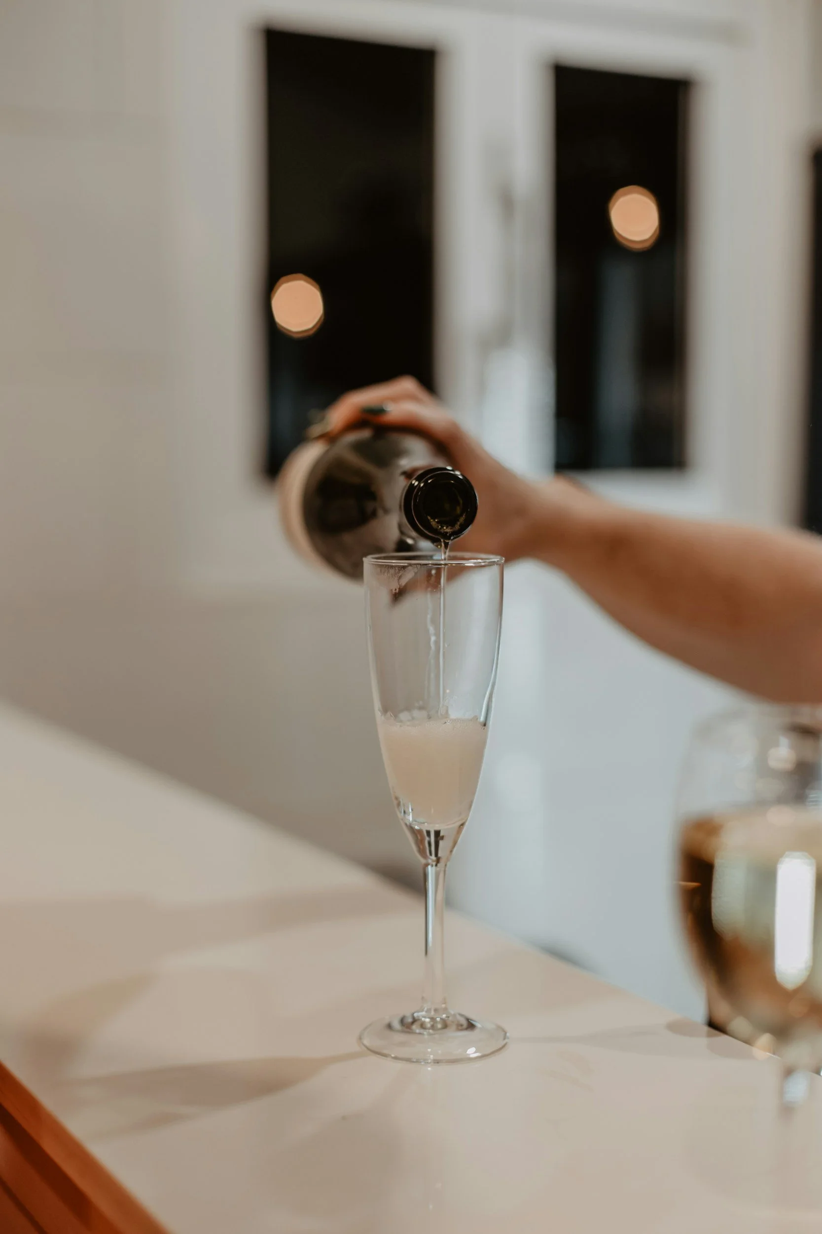 A person pouring champagne into a tall champagne flute at a bar or counter with a blurry background.