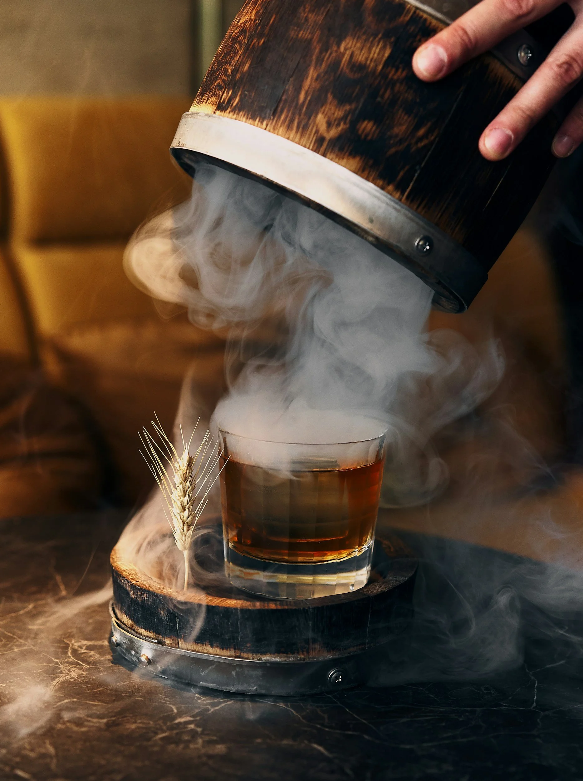A hand pouring dry ice into a glass of dark beverage, creating a fog effect, on a wooden coaster with a wheat stalk nearby.