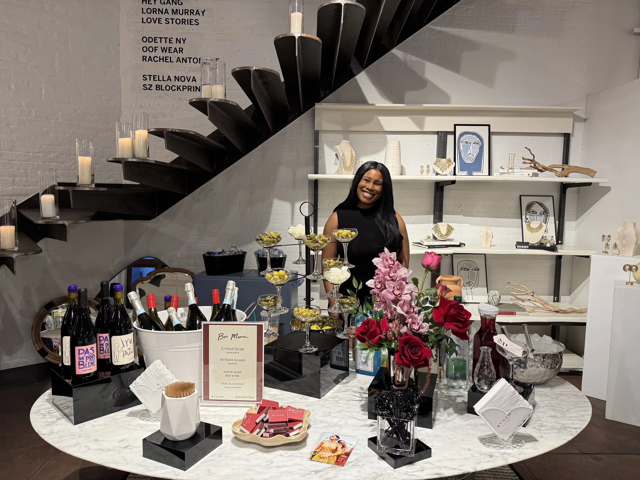 A smiling woman standing behind a table with wine bottles, flowers, and snacks in a boutique setting with artwork on the shelves and candles on a staircase.