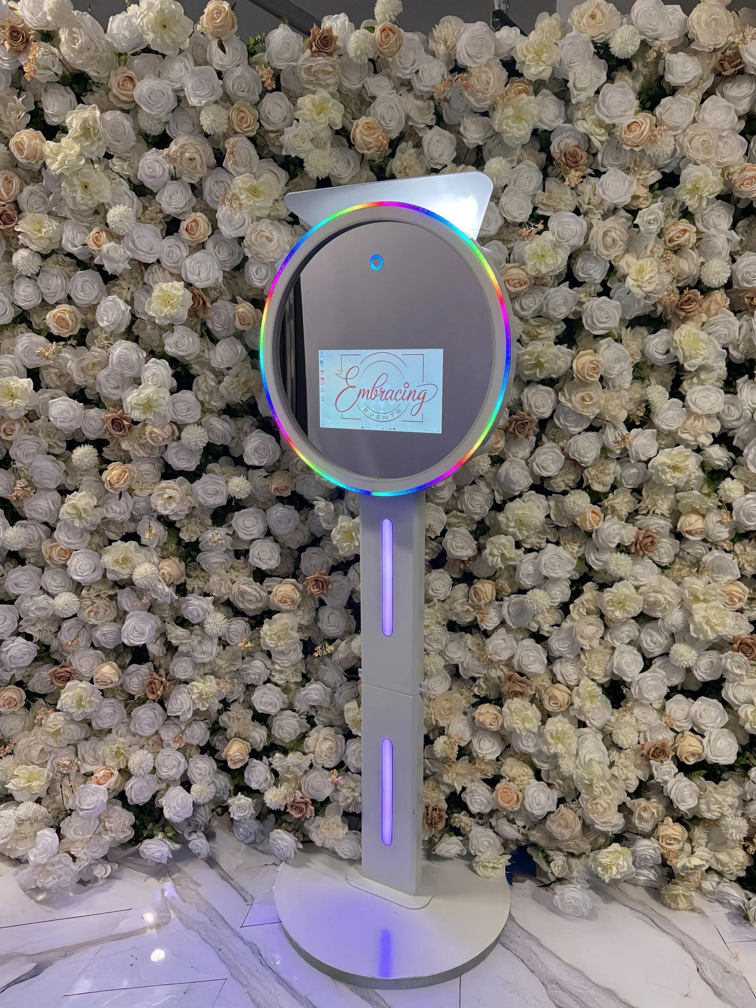 A digital photo booth with a circular mirror surrounded by rainbow LED lights, set against a backdrop of an arrangement of white and pale peach roses and cream-colored flowers, on a marble floor.