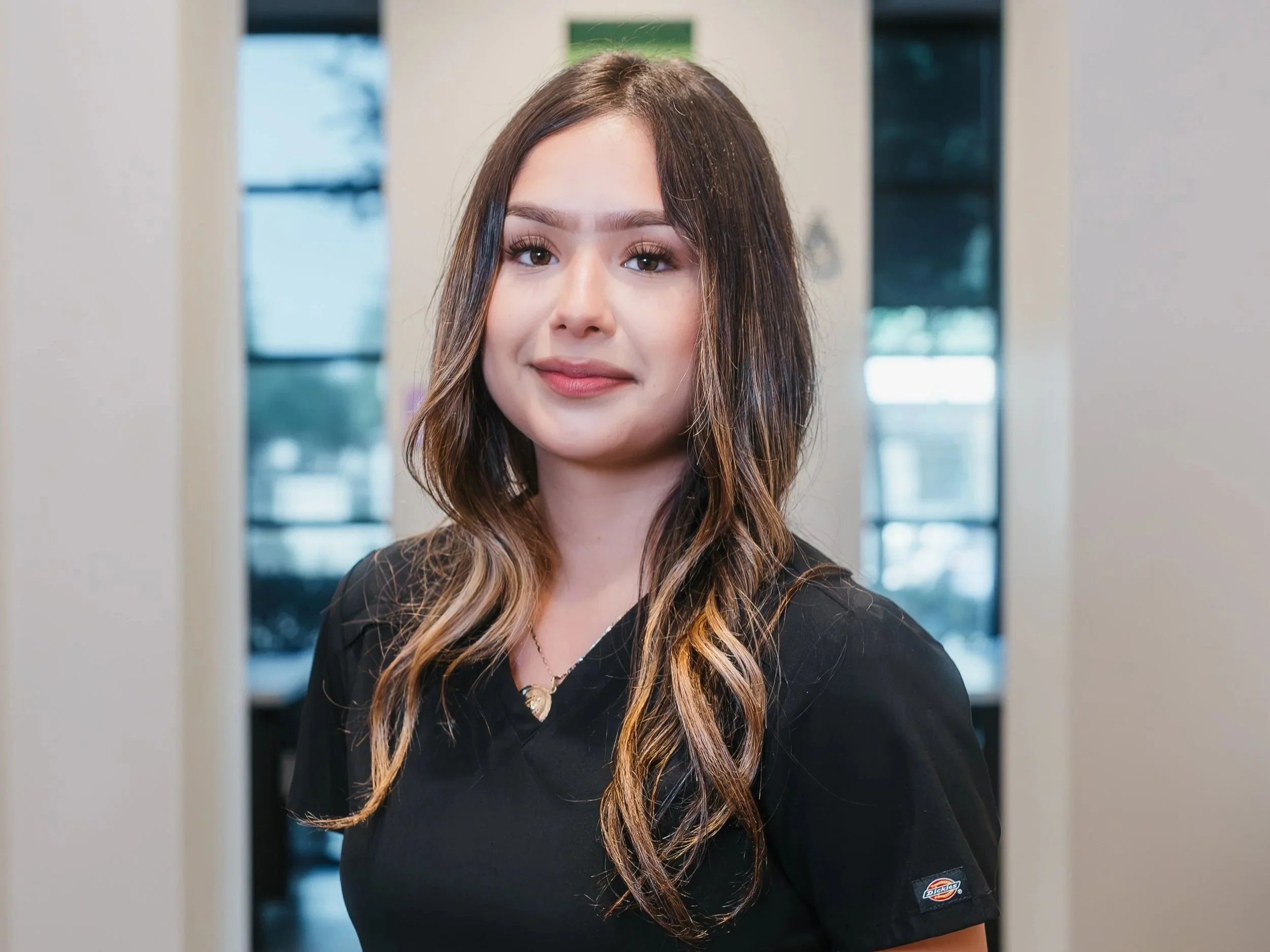 Woman smiling with long dark hair, wearing a black top, and a blurred outdoor background.