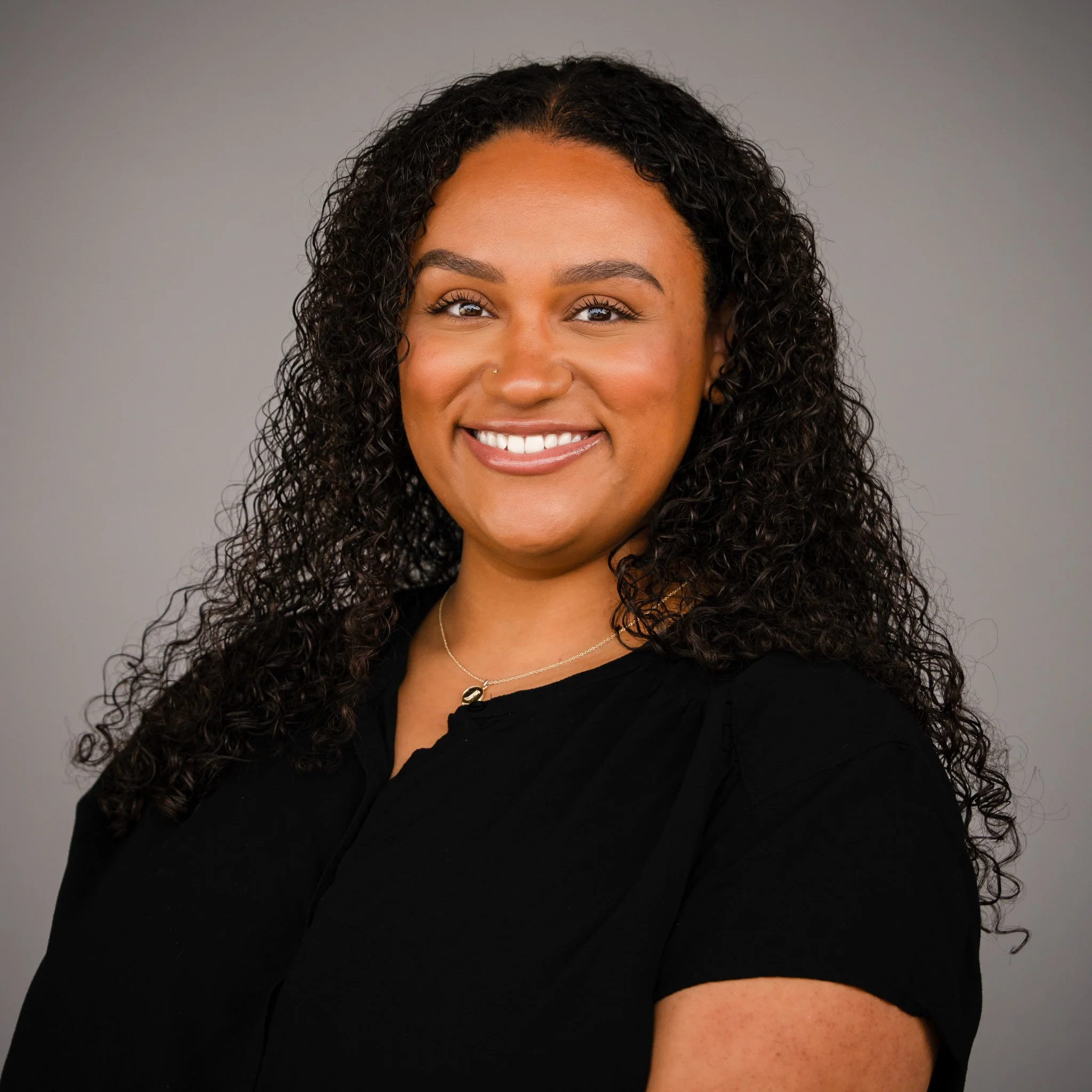 Smiling person with long dark hair and braces, wearing a dark-colored top, standing outdoors.