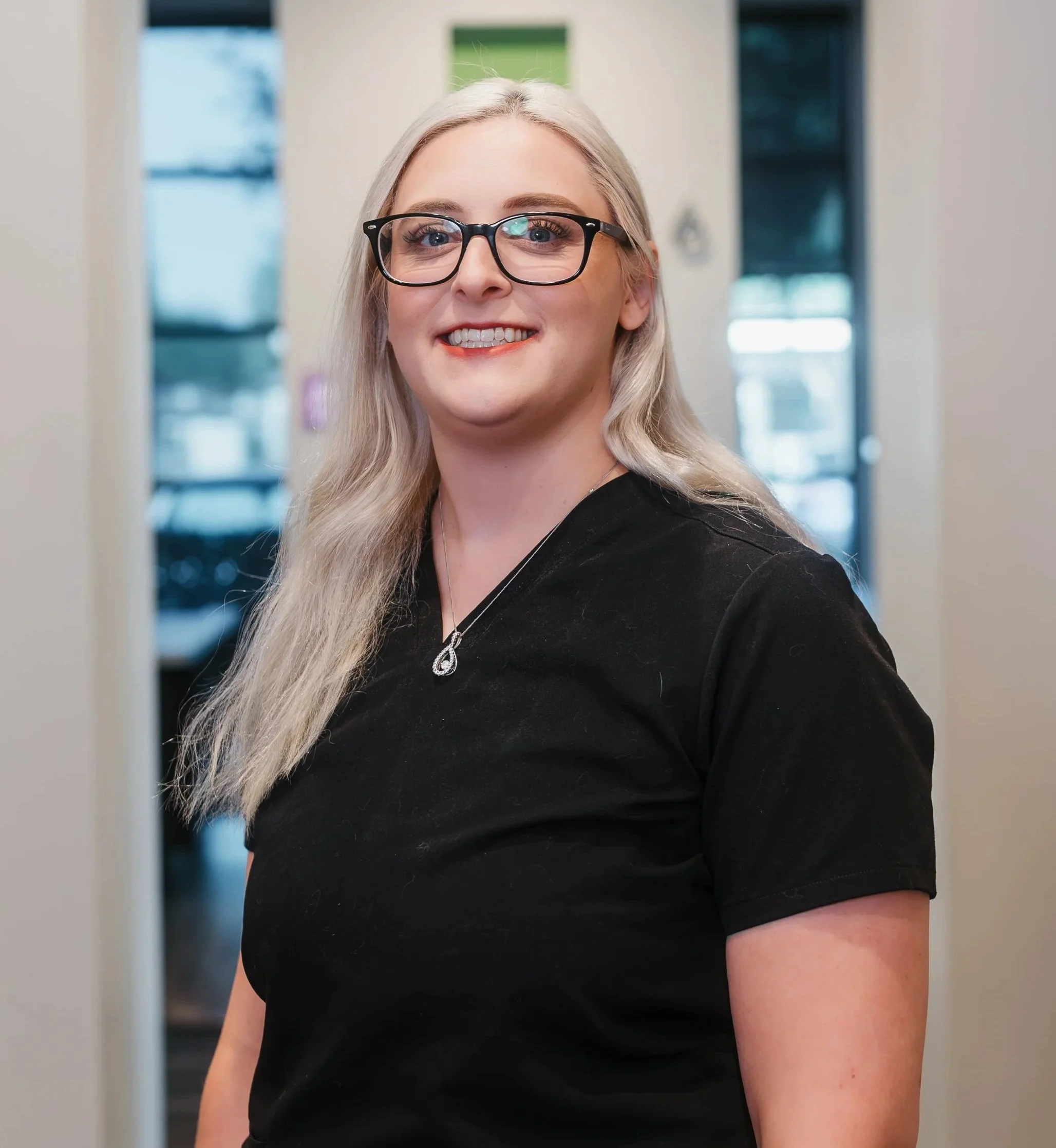 Person smiling with glasses and wearing a black top, standing in front of a brick wall.
