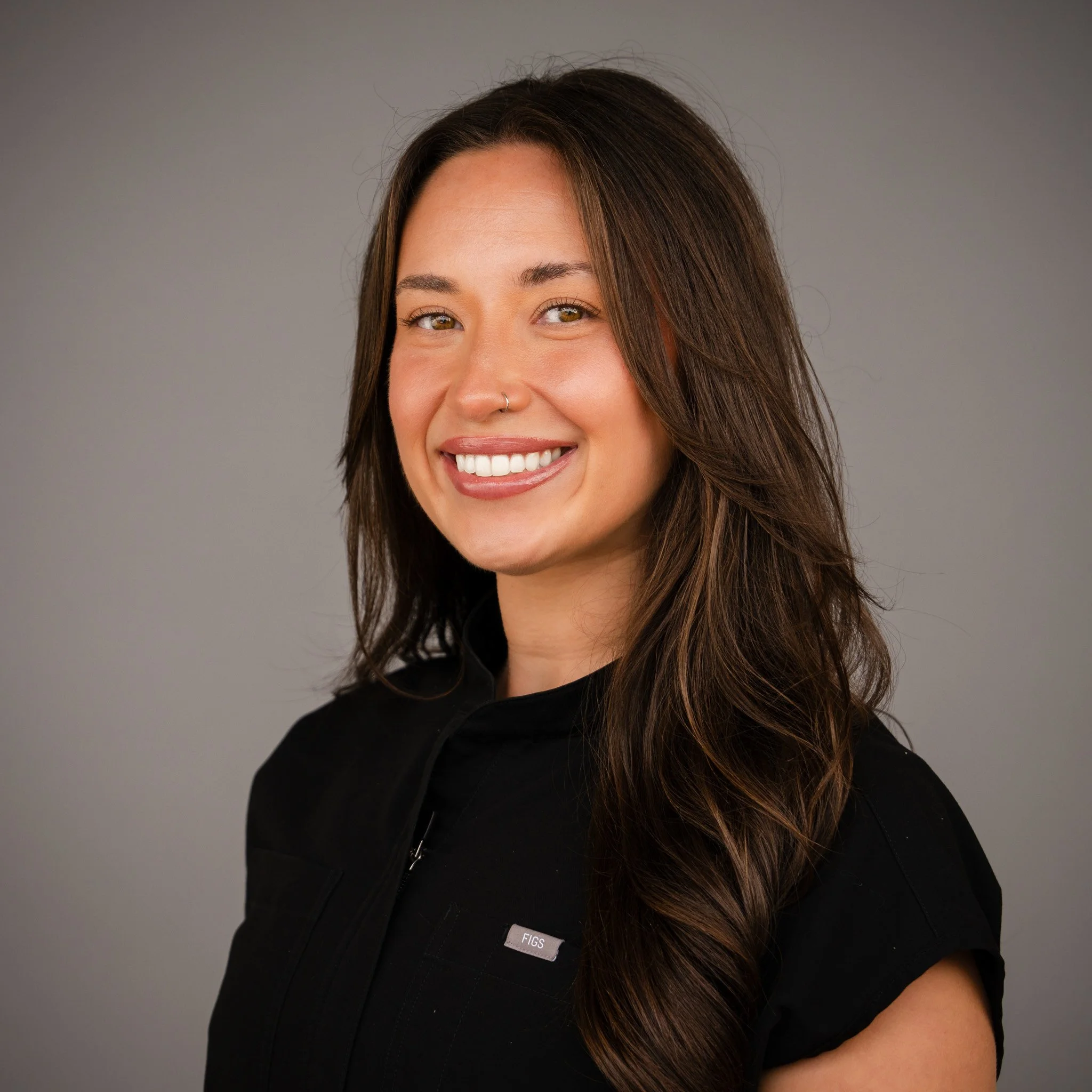 Smiling person with long dark hair and a nose ring, standing in front of a beige brick wall.