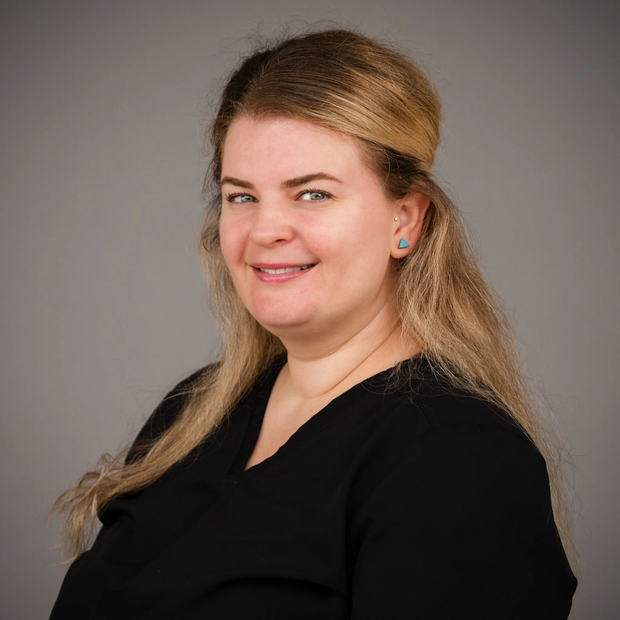 Woman with long blonde hair smiling, wearing a black top, standing in front of a beige brick wall.