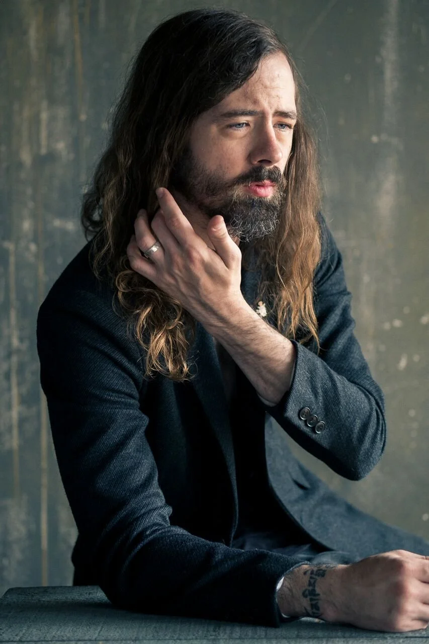 Man with long hair and beard in a suit, touching his face; tattoo visible on wrist.