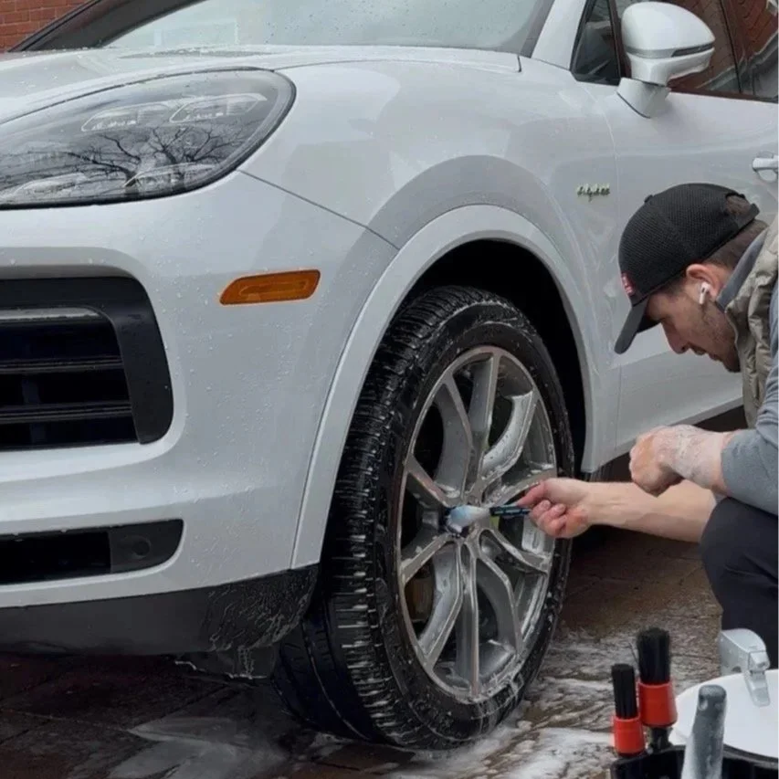 A man washing or detailing a silver SUV using a cleaning tool, with soap or water on the tire and on the ground.
