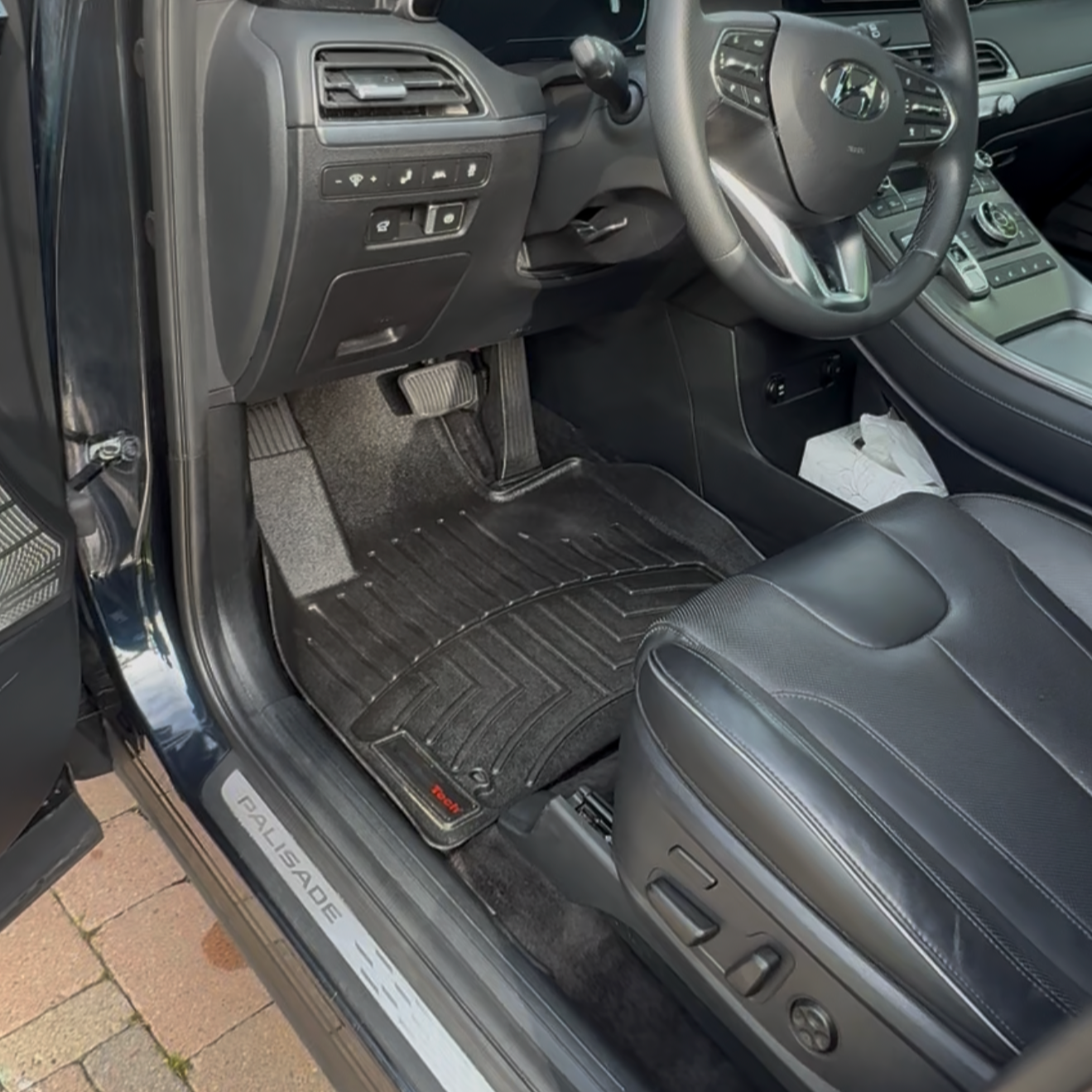 Inside a Honda Palisade SUV showing the driver’s side, including the steering wheel, dashboard, pedals, and a black leather seat with floor mats.