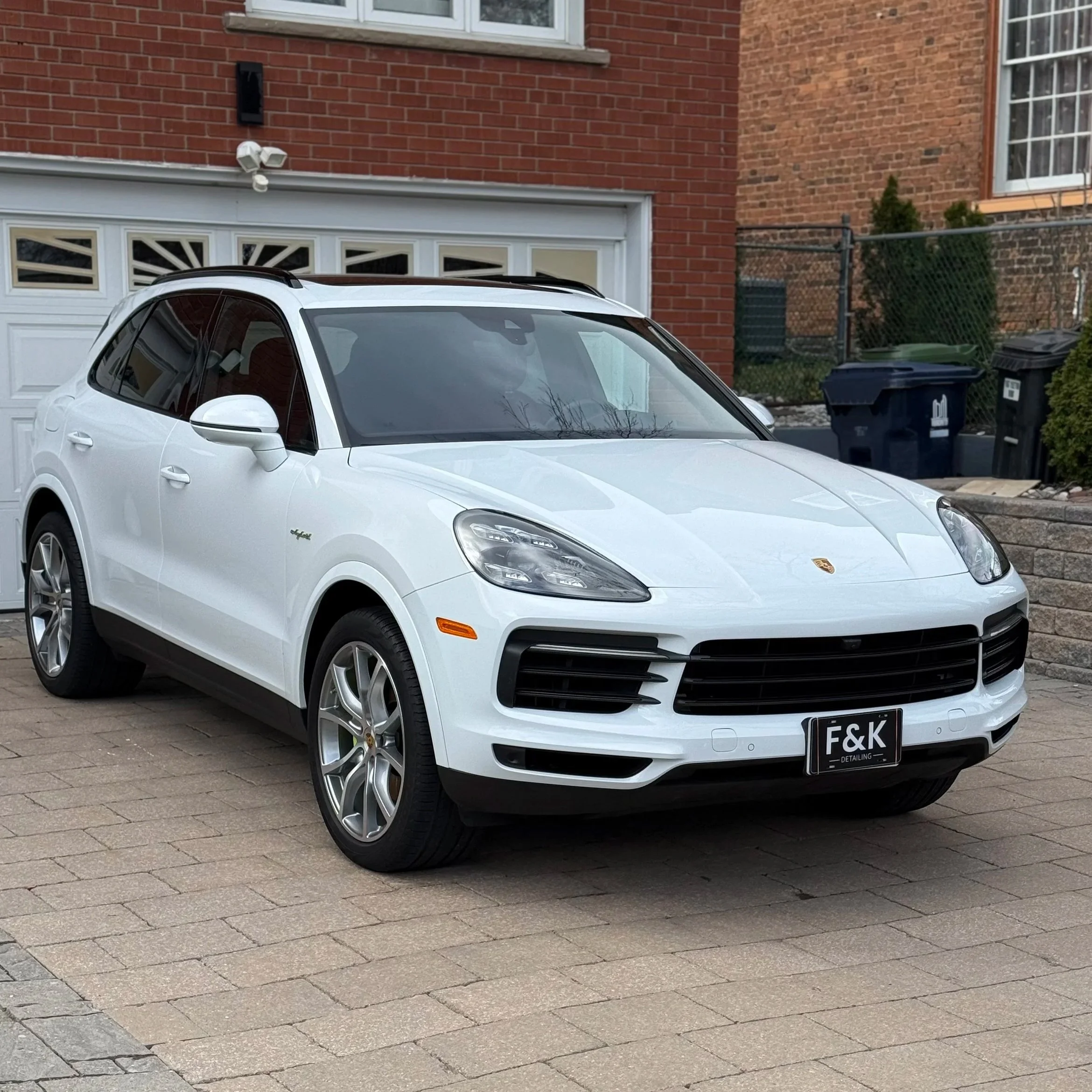 A white Porsche Macan parked in front of a house with a brick wall and a garage door. The car has black accents and a black license plate with the letters 'F&K'.