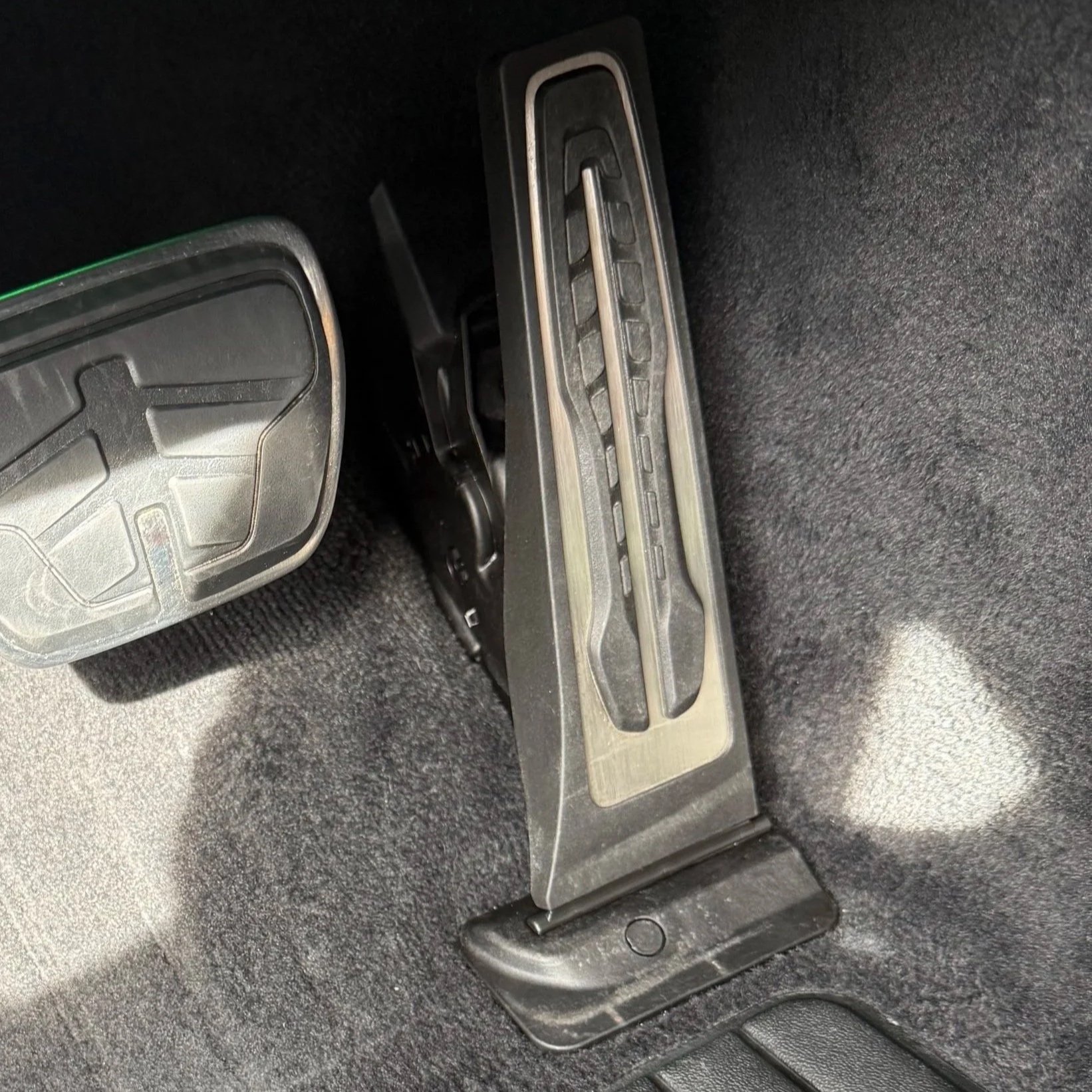 Close-up of car pedal set including accelerator, brake, and clutch pedals on a black textured floor mat.