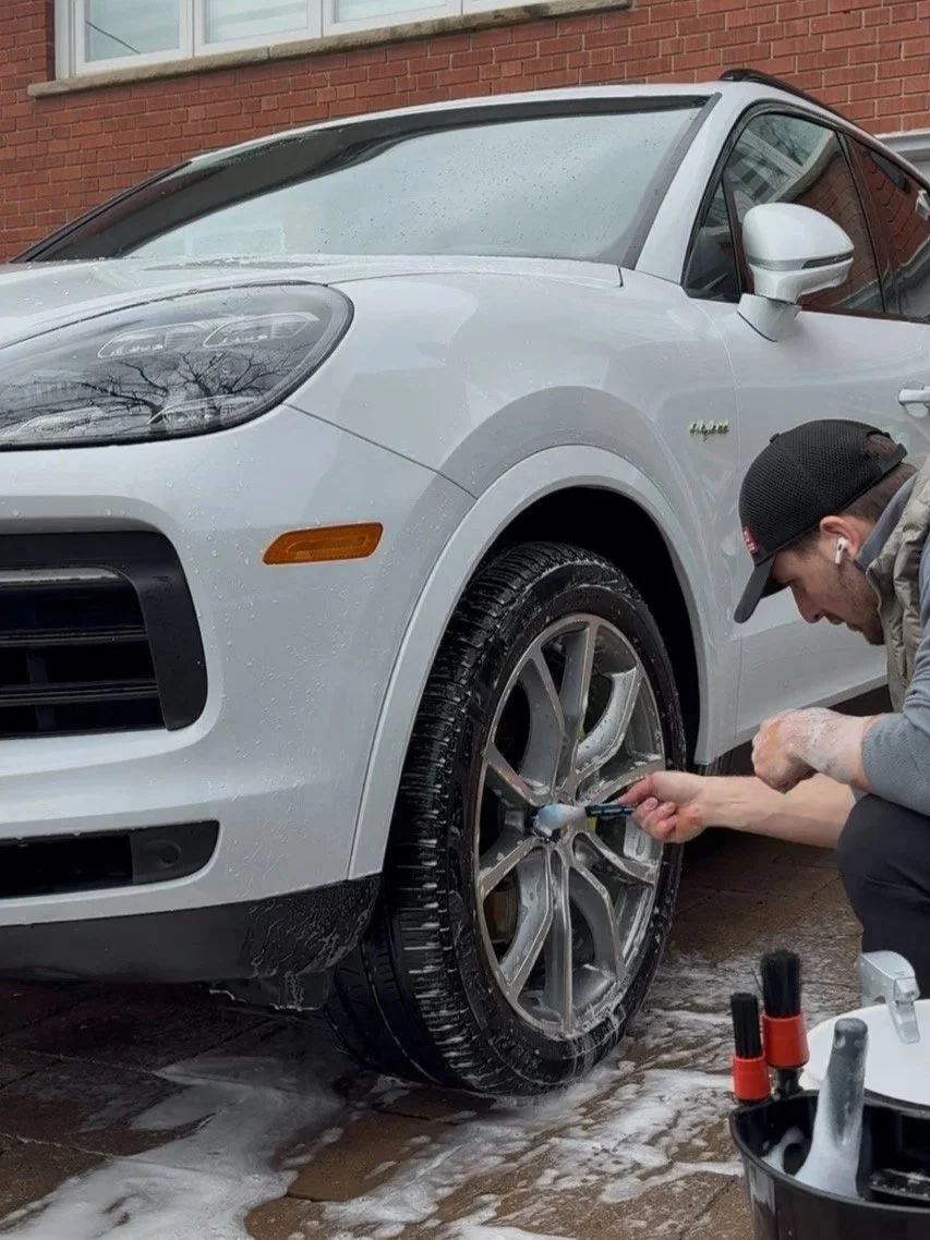 A man washing a white Porsche SUV with soap and water outside on a wet driveway.