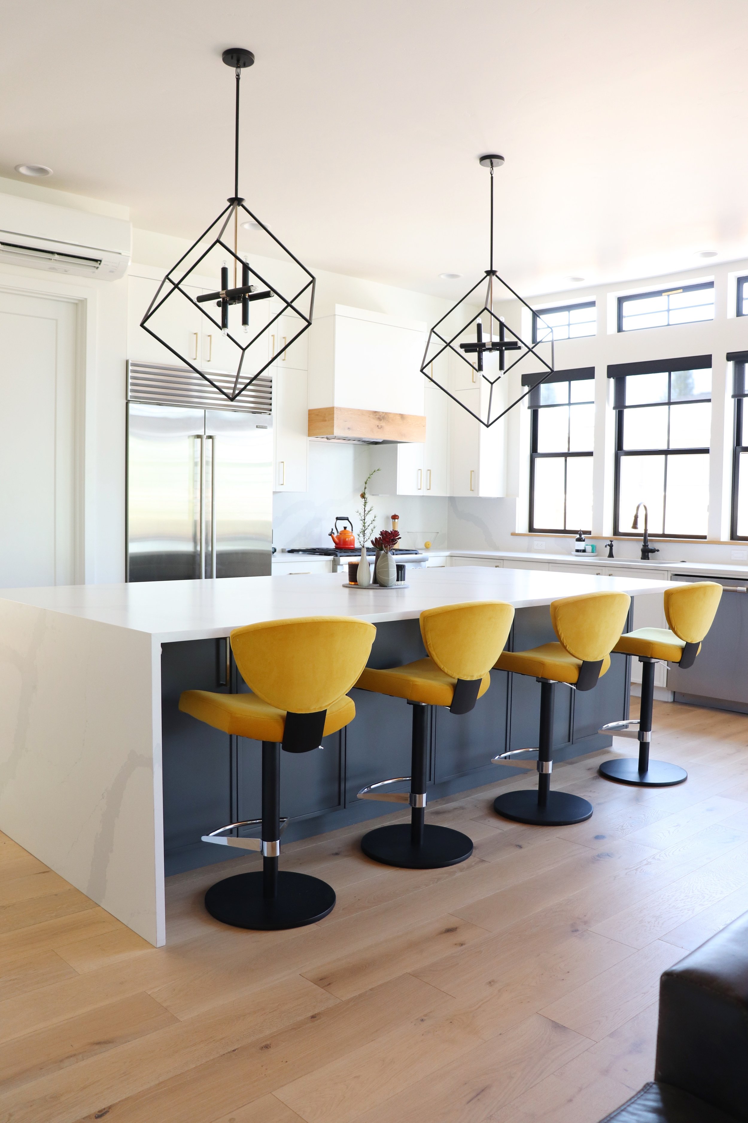 Modern kitchen interior design with white countertops, yellow bar stools, black pendant lights, wooden flooring, and large windows in Bend, Oregon.